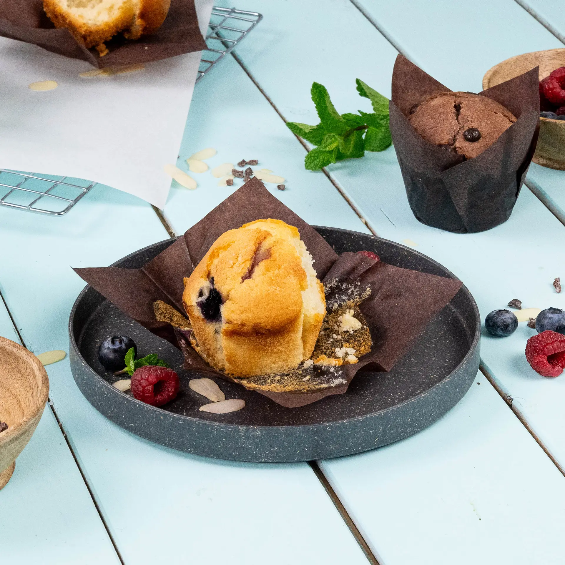Ein brauner Papier-Muffinbecher mit einem Blaubeermuffin auf einem dunklen Teller, umgeben von Himbeeren, Blaubeeren und einem Minzblatt.