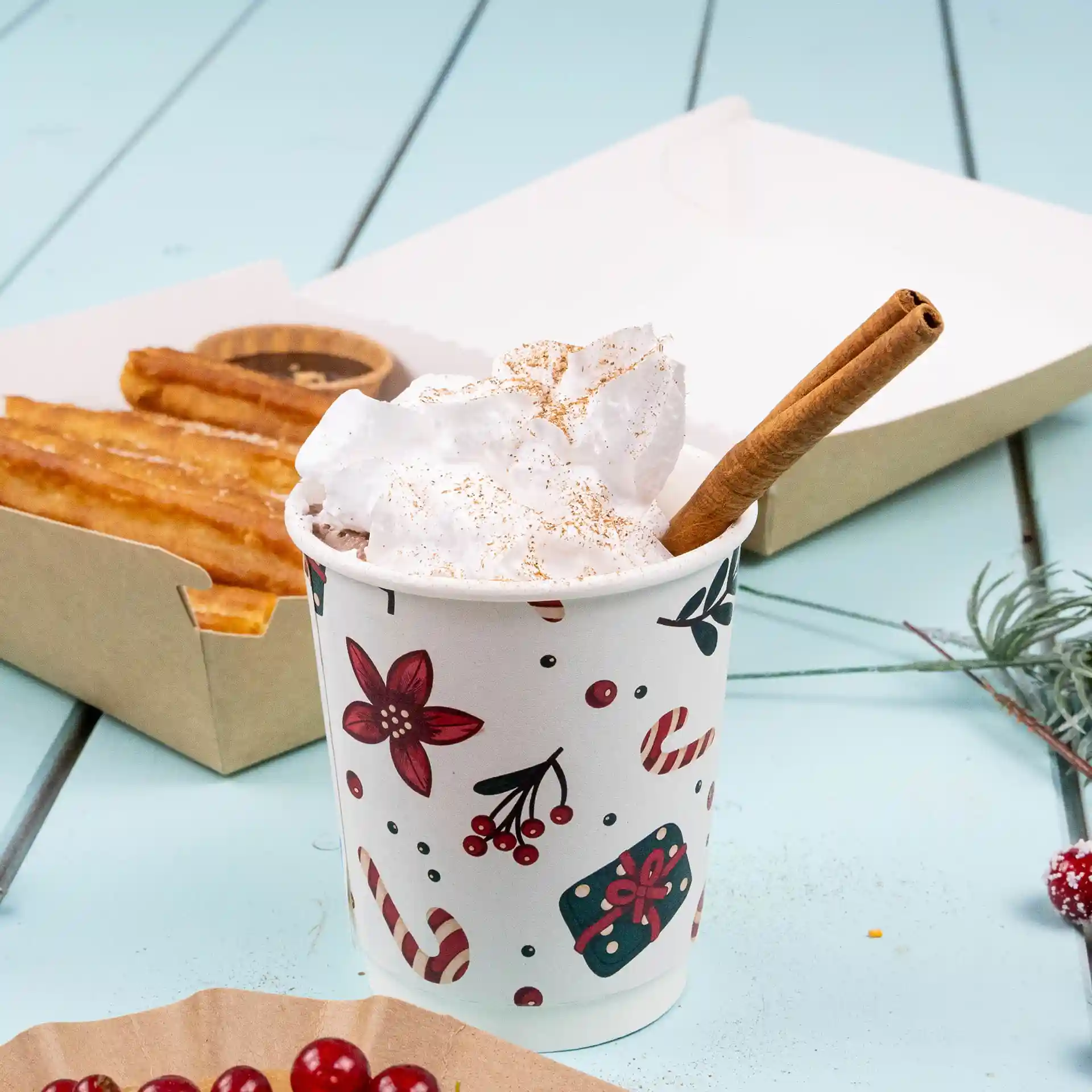 Doppelwandiger Becher mit Winterdesign, gefüllt mit Schlagsahne und einem Zimtstängel, neben Churros auf einer blauen Oberfläche.