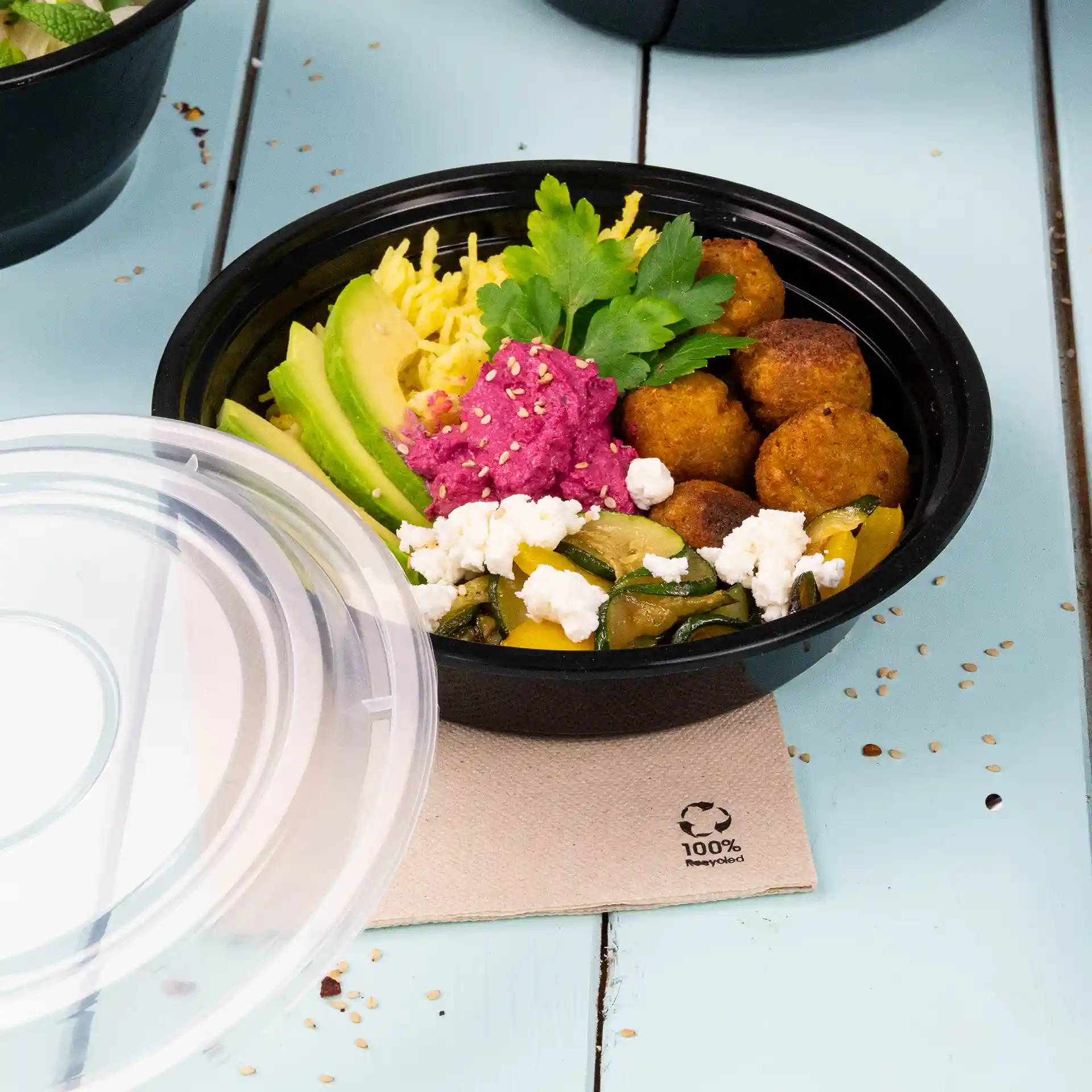 Piatto rotondo riutilizzabile nero con vari alimenti tra cui avocado, falafel e salsa rosa, parzialmente coperto da un coperchio trasparente.
