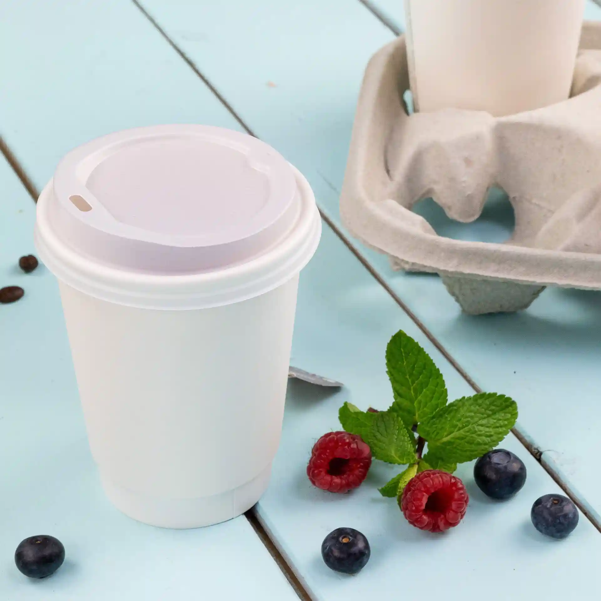 Tazza da caffè bianca a doppia parete con coperchio, 300 ml, posata su una superficie blu con lamponi, mirtilli e foglie di menta.