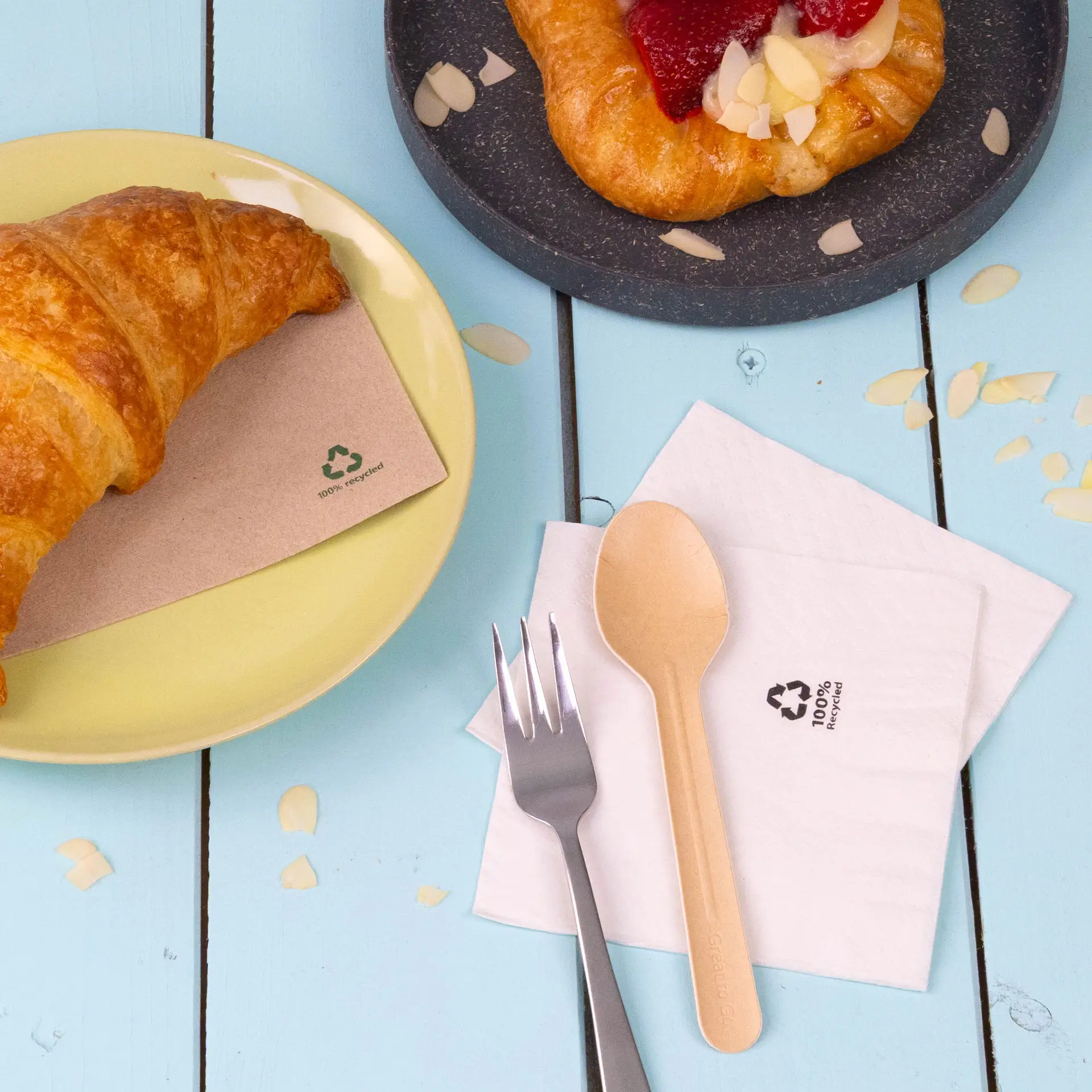 Ein Croissant auf einem gelben Teller neben einer weißen 2-lagigen, recycelten Papierserviette und einem umweltfreundlichen Holzlöffel und -gabel.