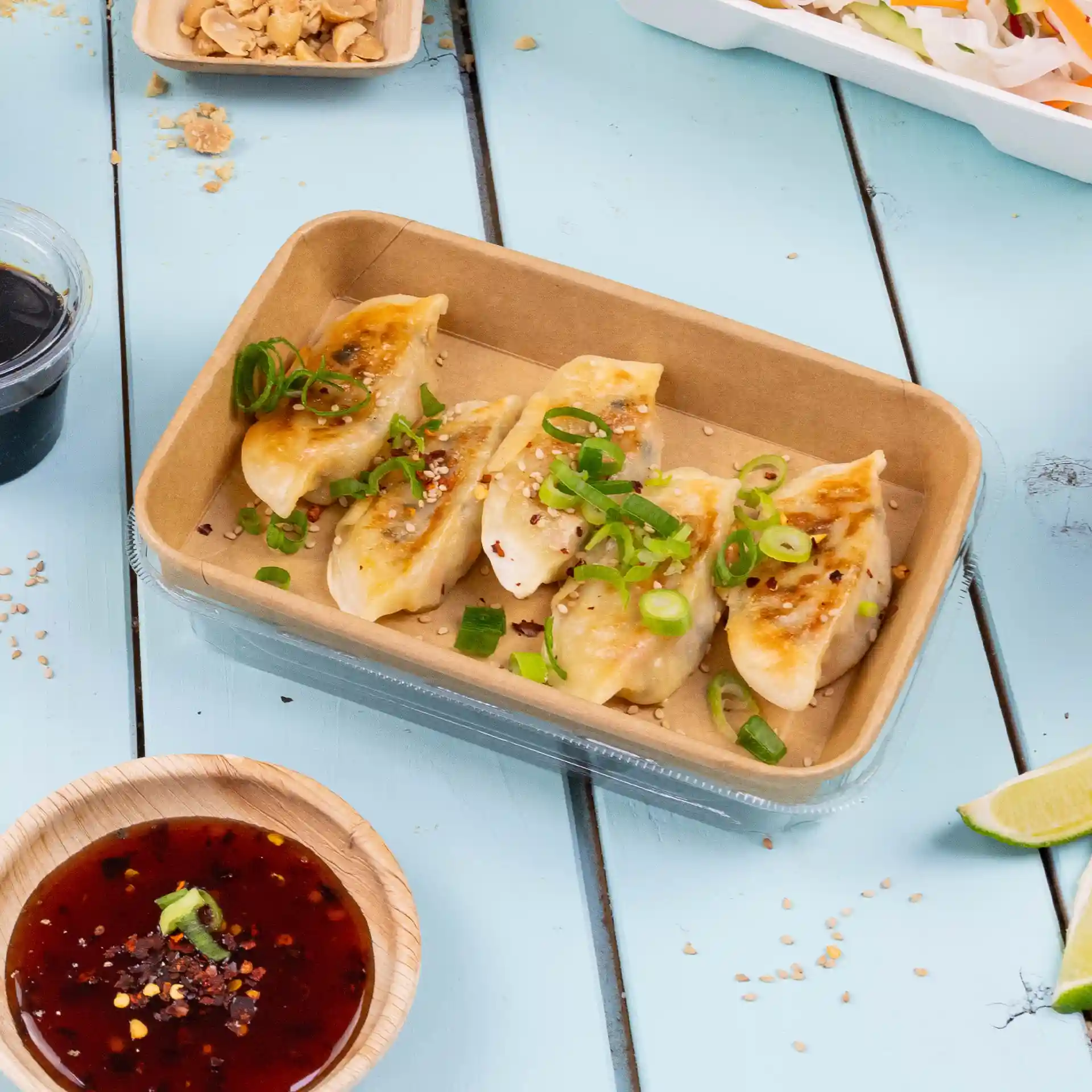 Behälter mit fünf Teigtaschen, garniert mit Frühlingszwiebeln und Sesam, serviert mit einer Dip-Sauce in einer Holzschale.