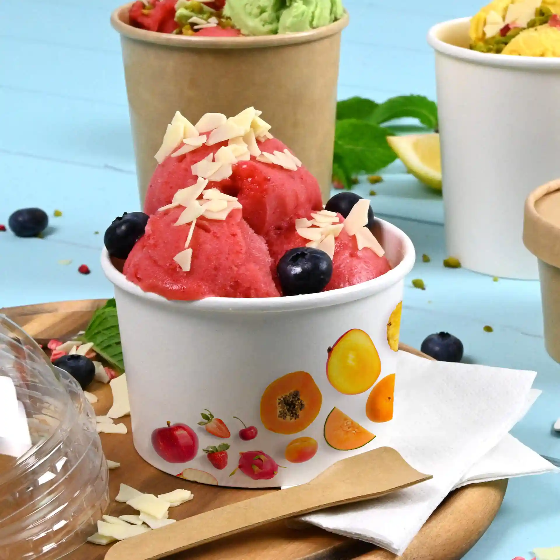 Runder weißer Becher mit rosa Sorbet, garniert mit Mandelstückchen und Heidelbeeren, auf einem Holztablett.