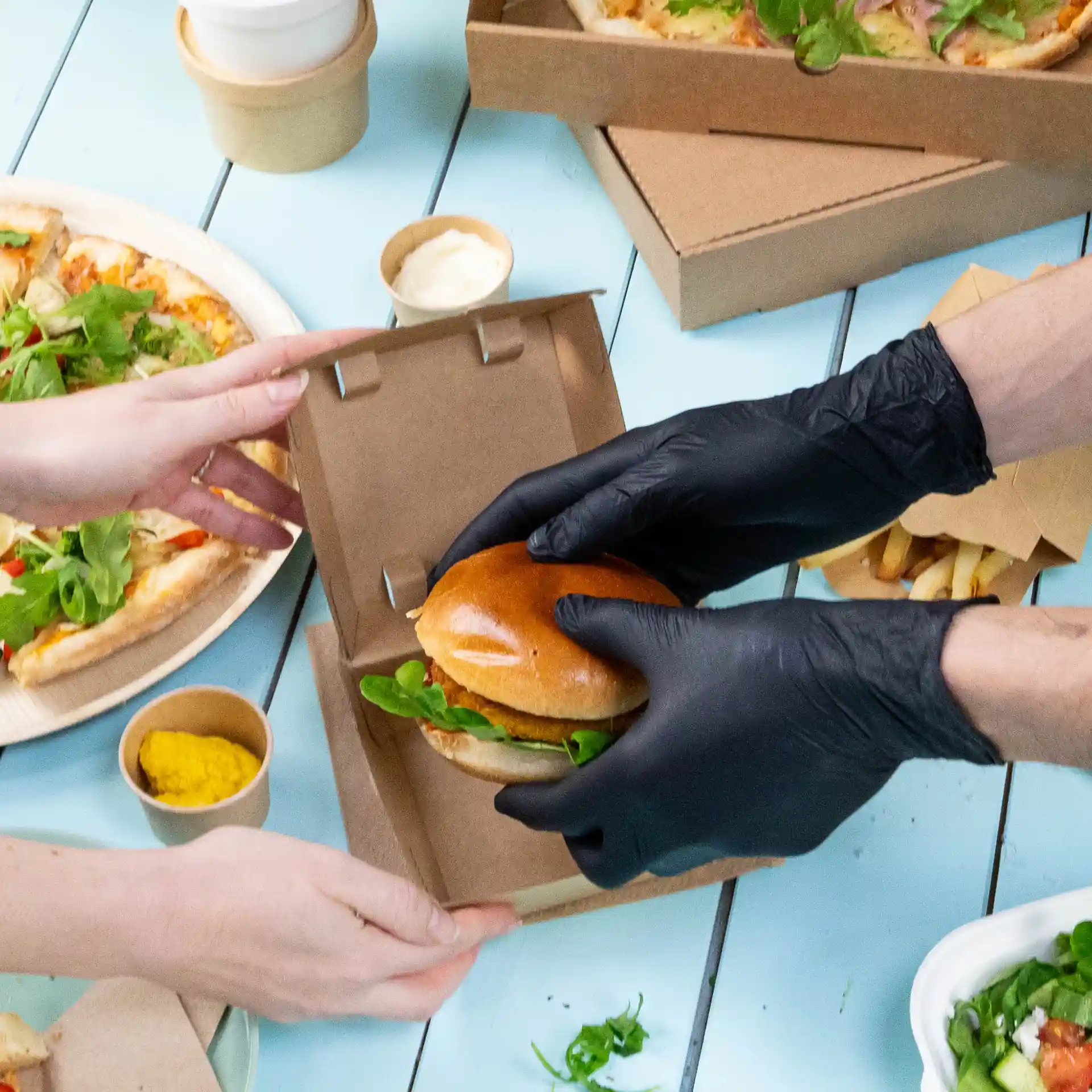 Image d'une personne portant des gants en nitrile noirs remettant un burger à une autre personne, avec des pizzas et des frites en arrière-plan.