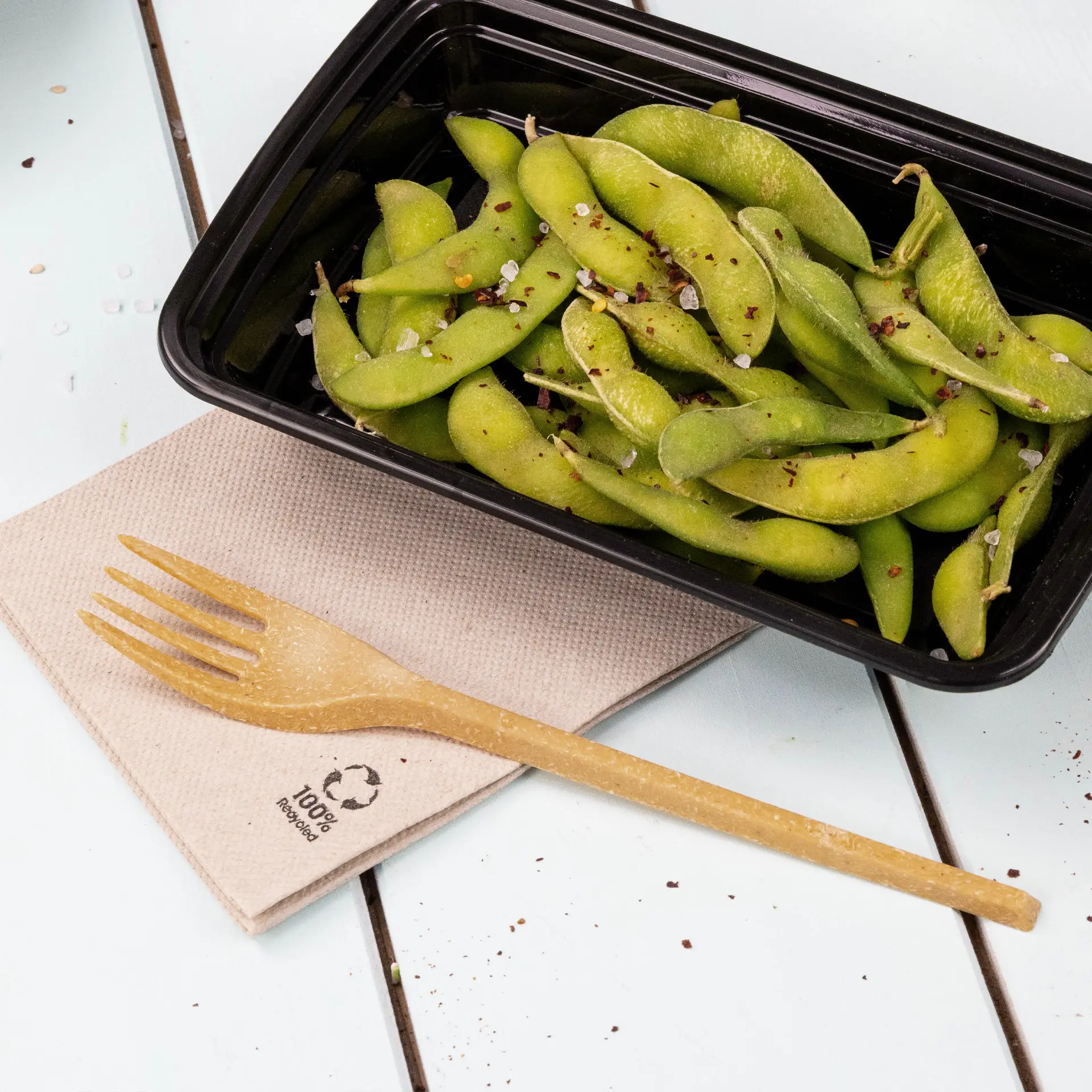 Ein schwarzer Behälter gefüllt mit grünen Edamame, garniert mit Gewürzen, begleitet von einer biologisch abbaubaren Gabel auf einer Recycling-Papierserviette.
