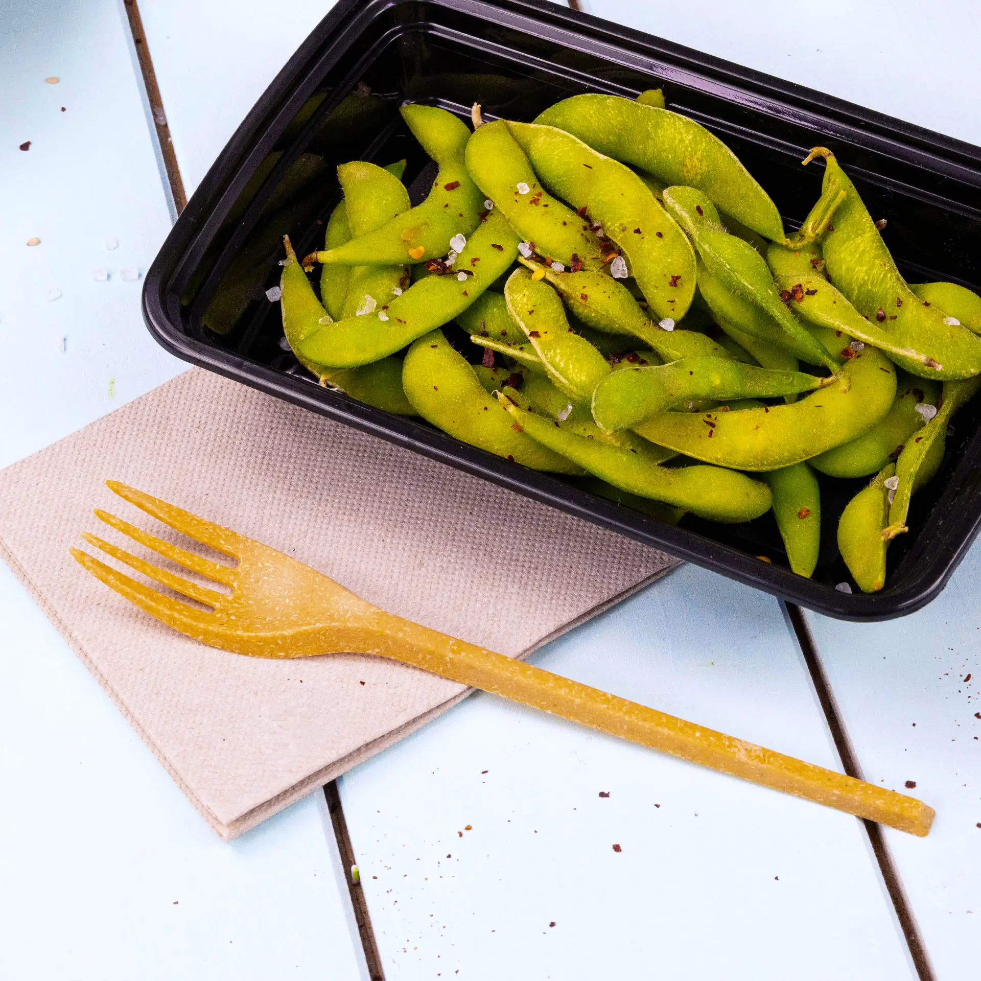 Bild eines schwarzen Behälters mit grünen Edamame-Bohnen, eine gelbe Gabel auf einer beigen Serviette daneben.