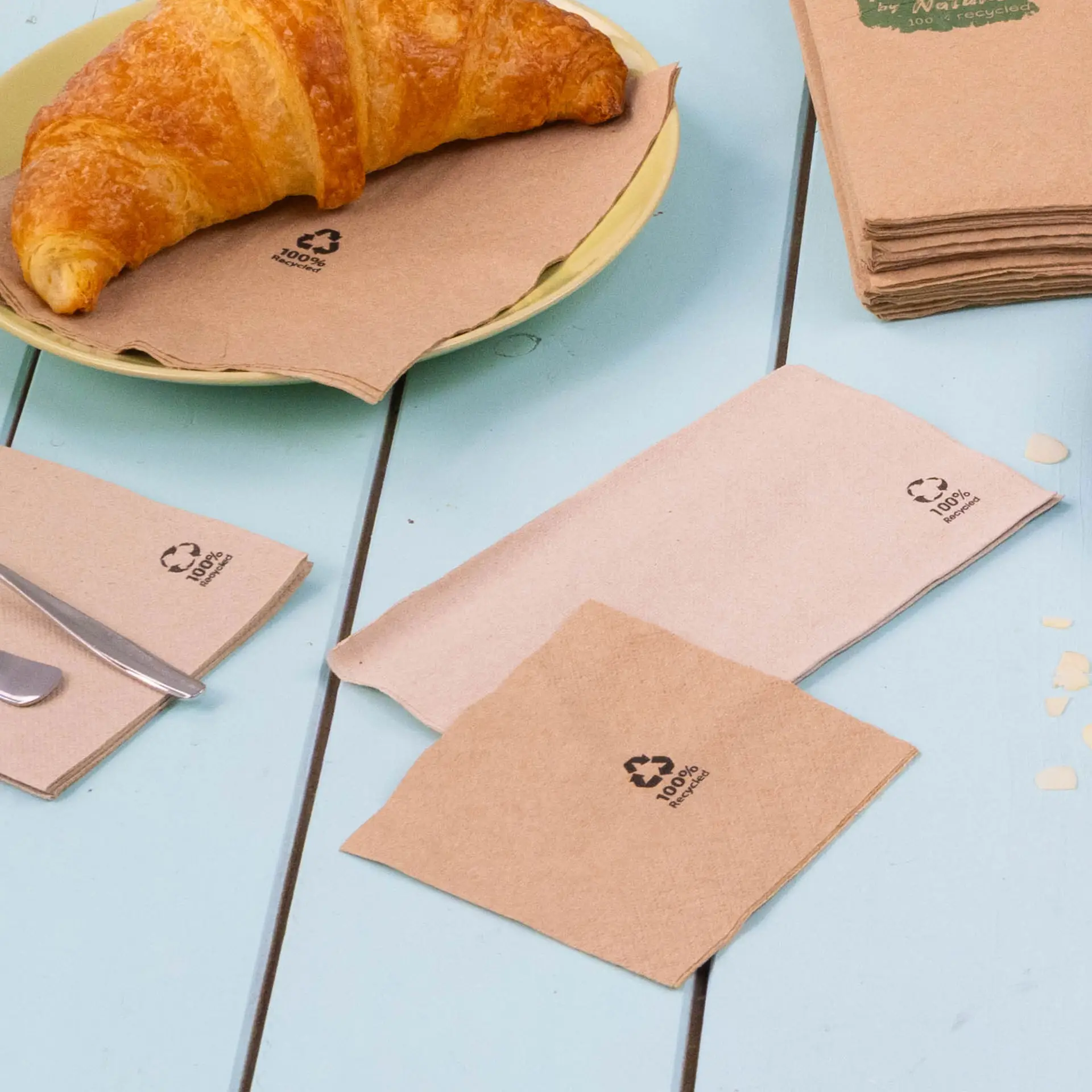 Image de serviettes à cocktail biodégradables marron sur une table bleue, avec un croissant sur une assiette, montrant des symboles de recyclage.