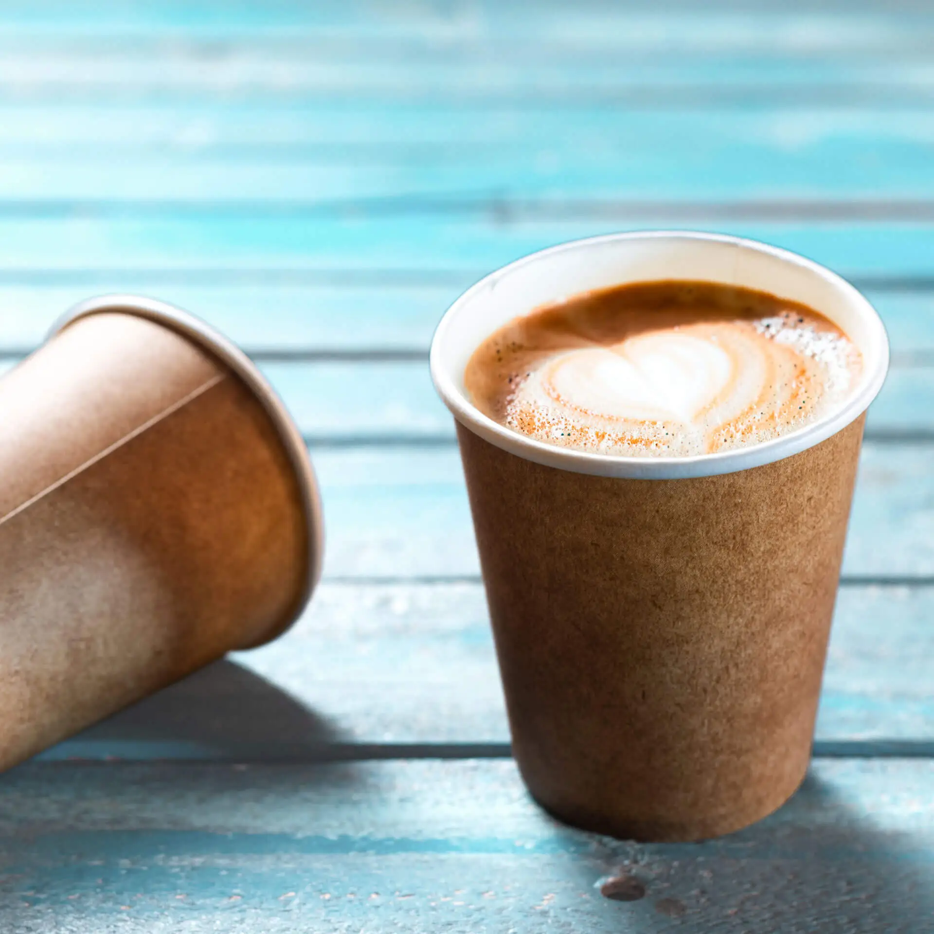 Bicchiere di carta marrone con interno bianco, riempito di caffè e arte latte a forma di cuore, su una superficie in legno blu.