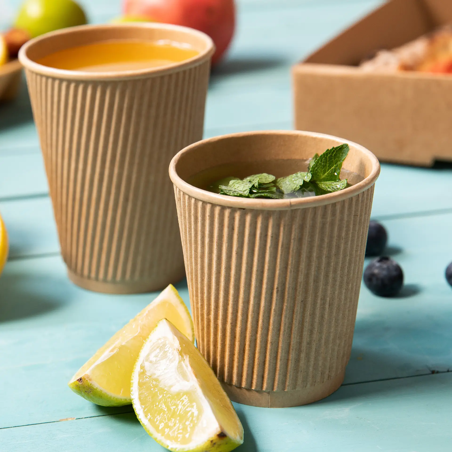 Deux gobelets en papier bruns, à rayures, sur une table bleue, l'un rempli de thé et de feuilles de menthe, l'autre de jus, à côté de quartiers de citron.