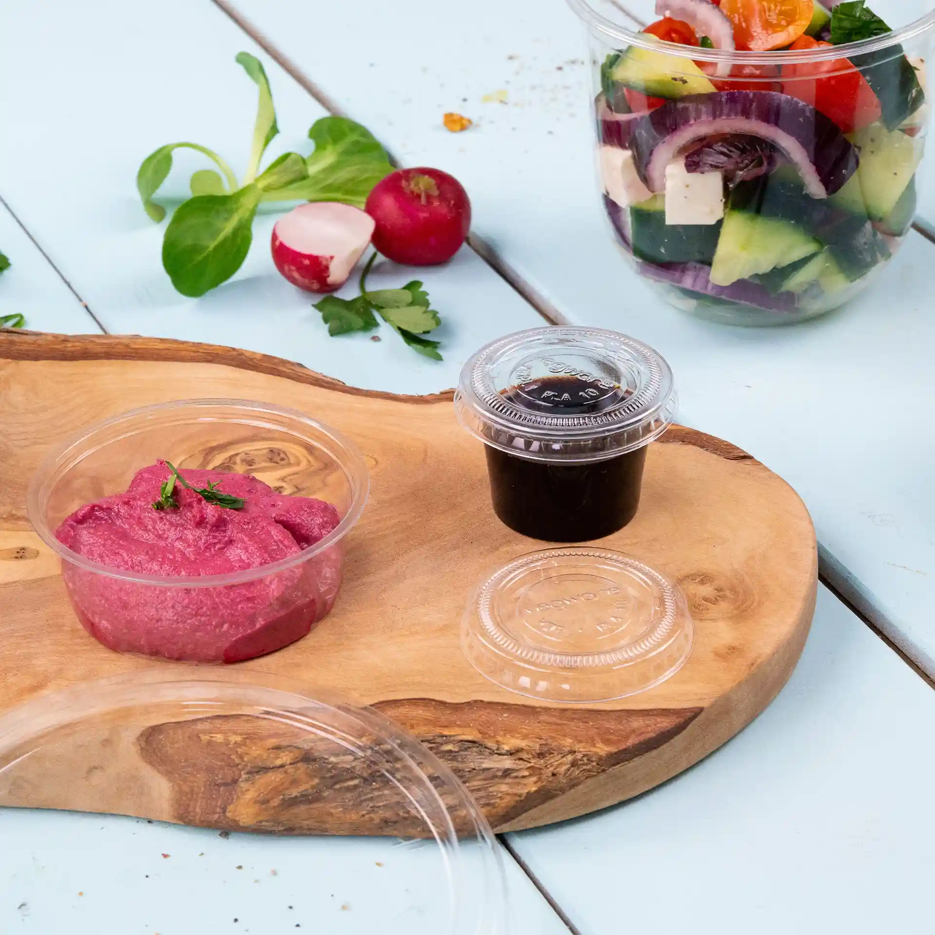 Immagine di un tagliere di legno con una crema rosa in un bicchiere trasparente, una salsa scura in un altro bicchiere trasparente e verdure fresche sullo sfondo.