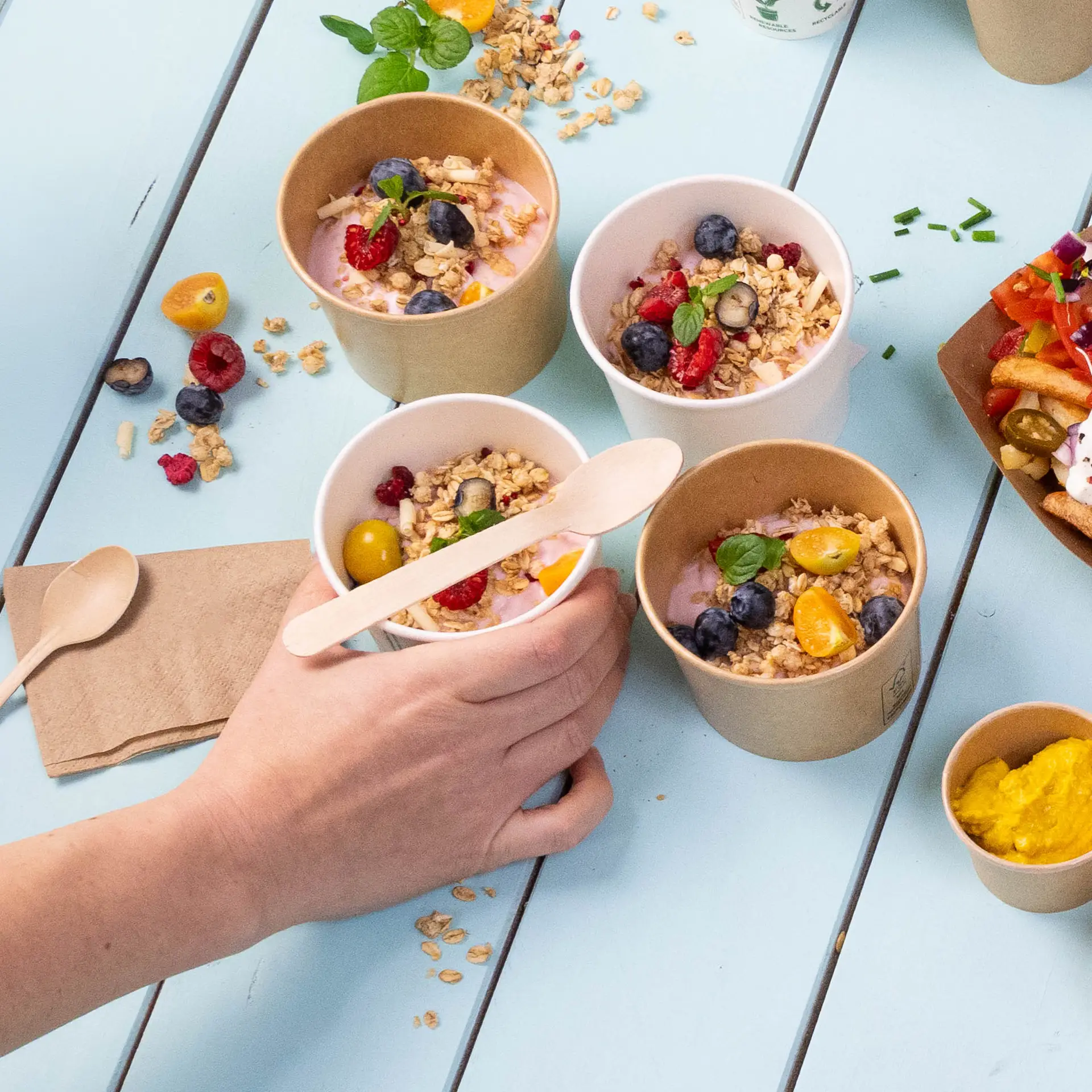 Eine Hand hält einen kleinen Holzlöffel über mehreren Bechern Joghurt, die mit Granola und Obst auf einem blauen Tisch belegt sind.