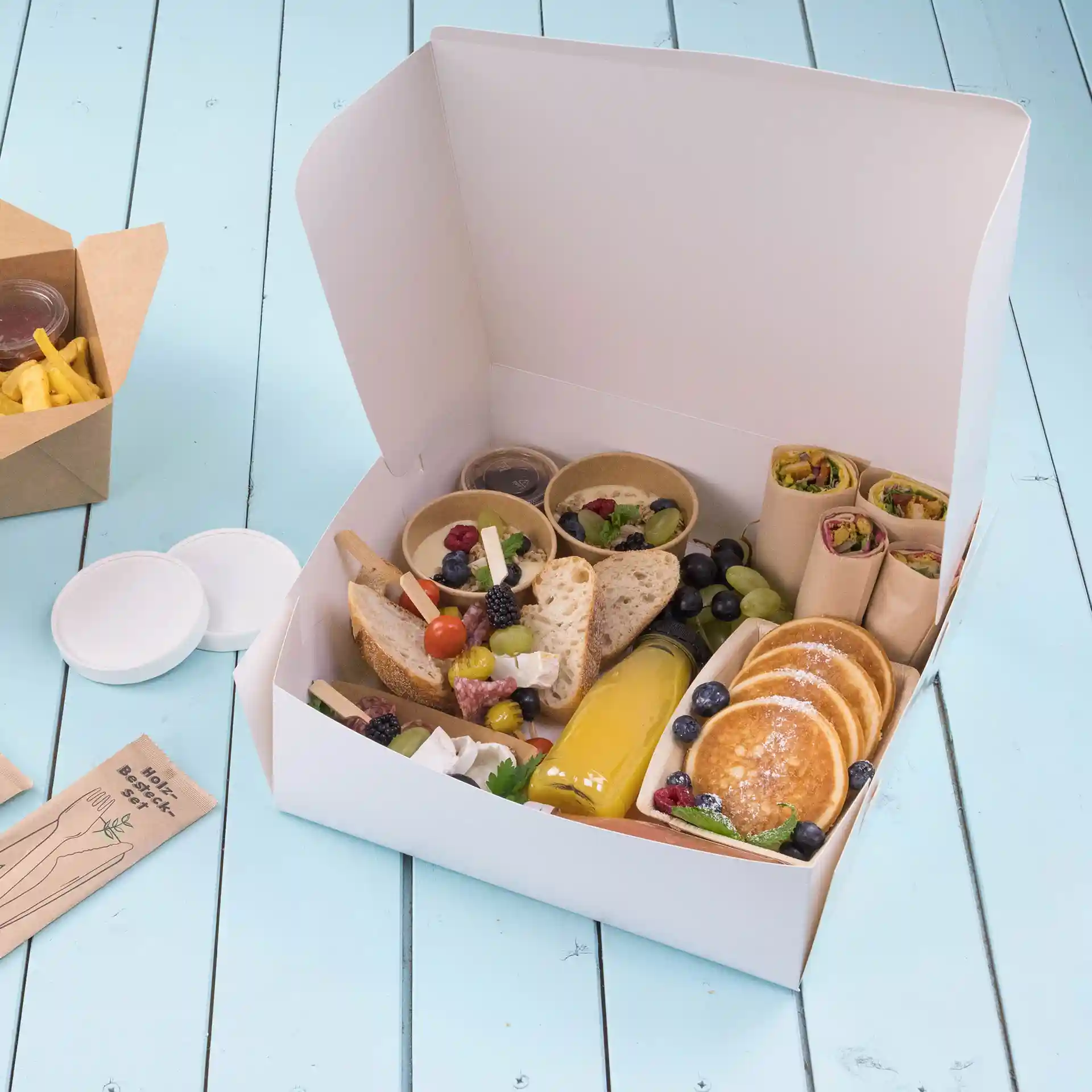 Boîte à gâteau blanche remplie d'aliments variés, y compris du pain, des fruits, des crêpes et des sauces, sur une surface en bois bleu.