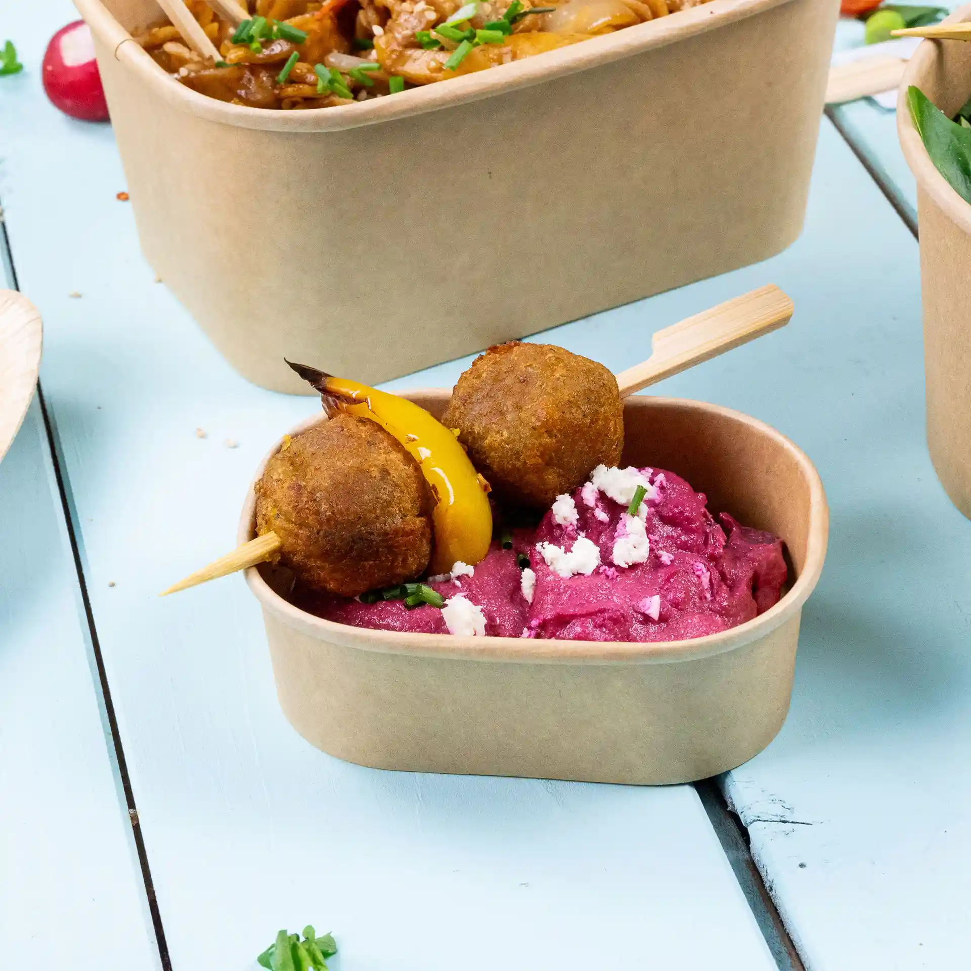 Contenitore alimentare rettangolare in kraft con hummus rosa e due polpette infilzate, guarnite con peperone giallo.