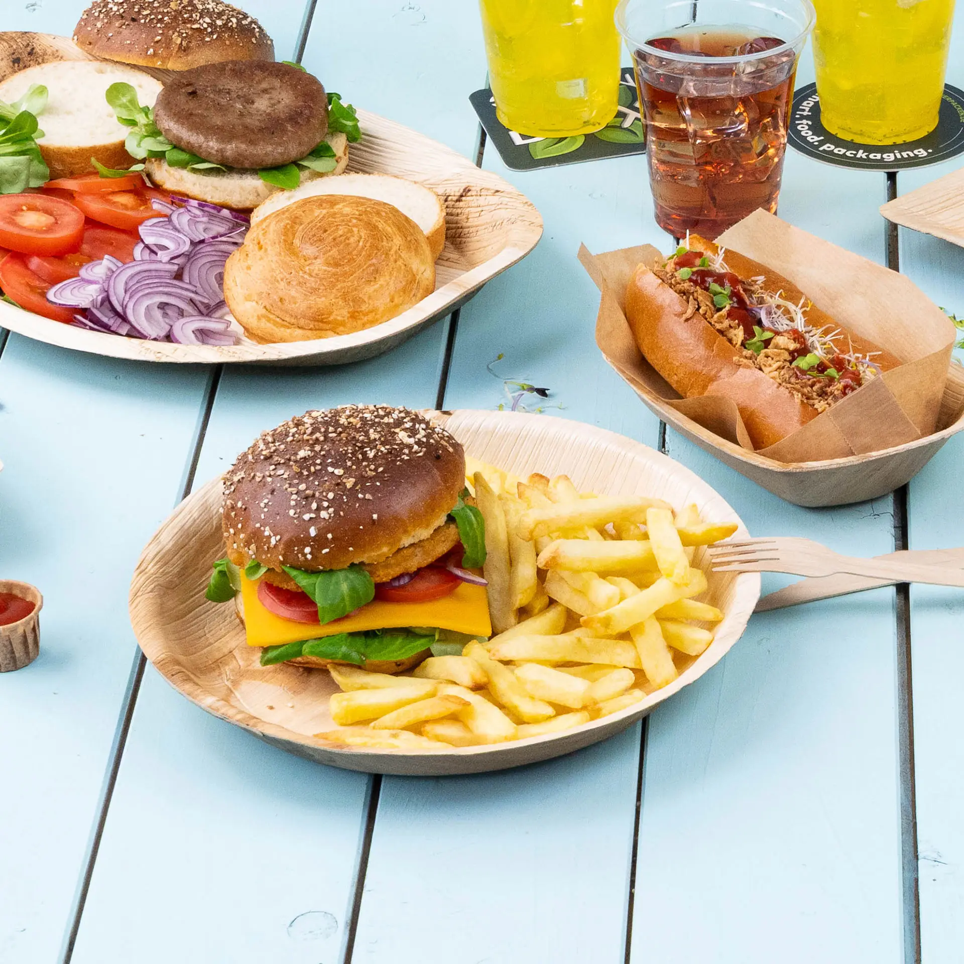 Runder Teller aus Palmblatt mit einem Hamburger, belegt mit Käse, Salat und Tomate, serviert mit Pommes auf einem blauen Tisch.