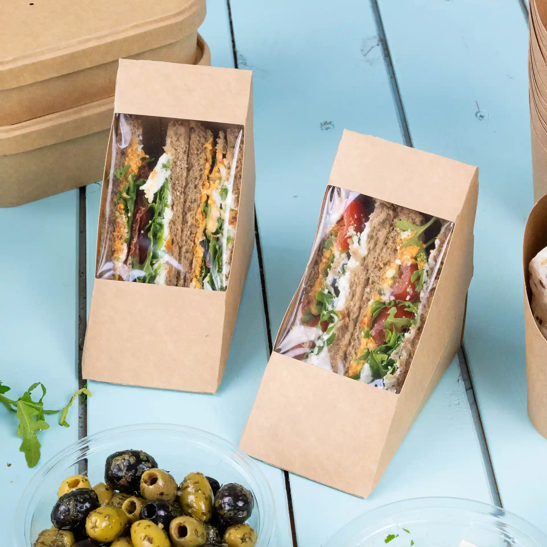 Deux triangles de sandwich en carton avec une fenêtre transparente, garnis de légumes, sur une surface bleue.