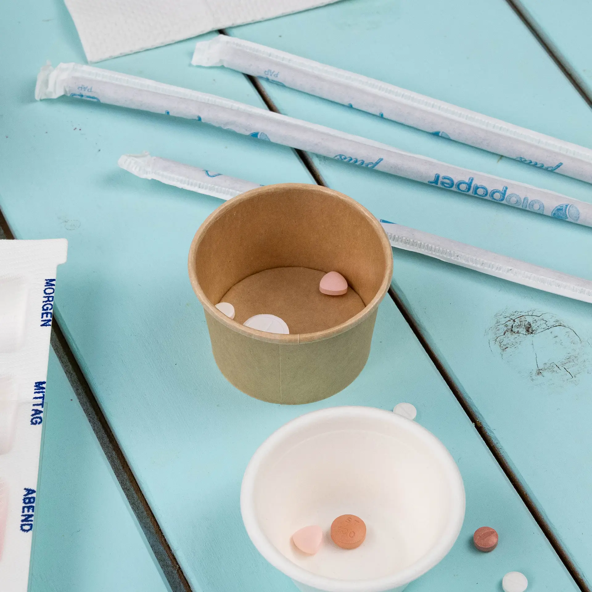 Deli-Container (braun, 40 ml) mit kleinen bunten Pillen; zusätzliche Behälter und Strohhalme liegen auf einer blauen Oberfläche.