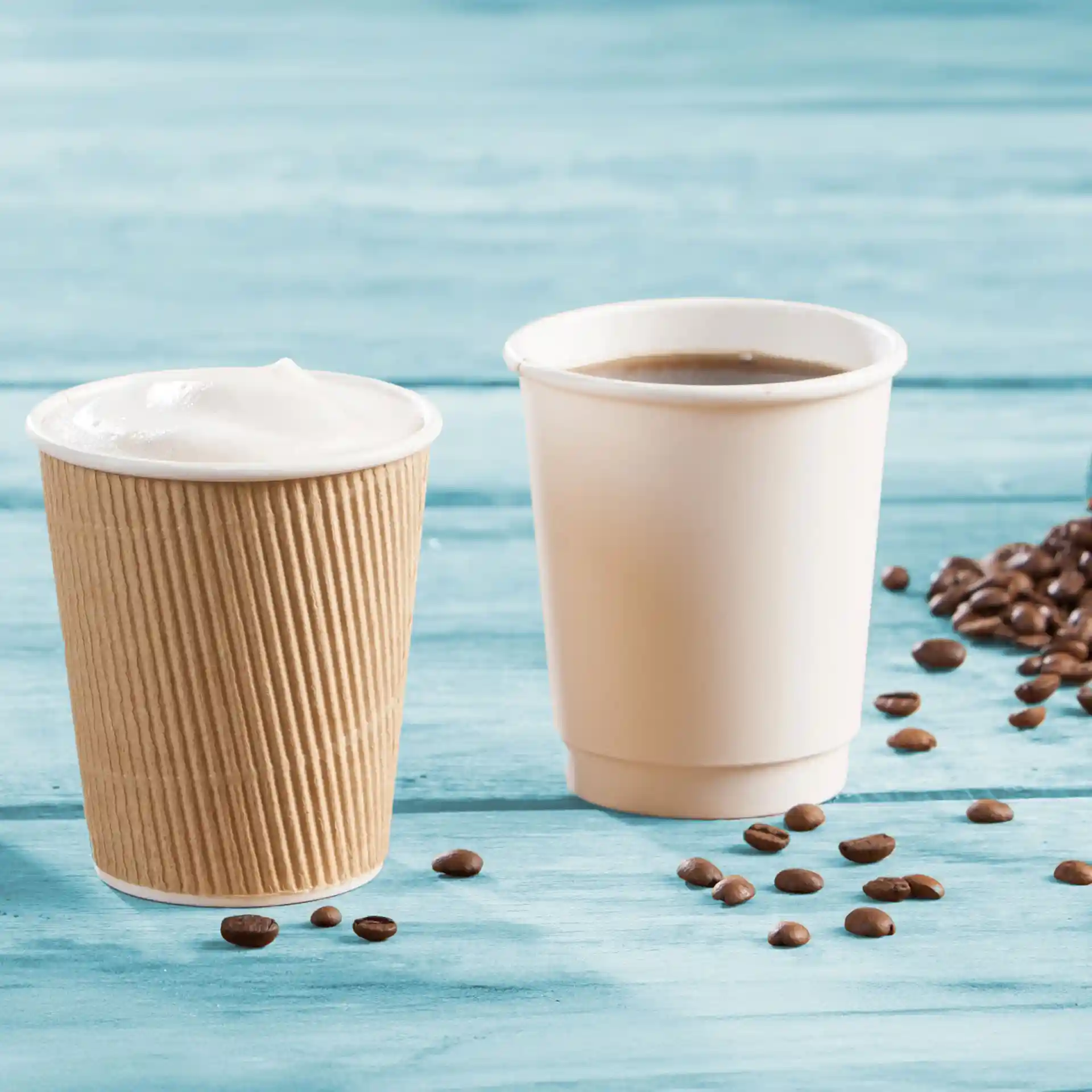 Bicchieri di cartone a doppia parete, uno con schiuma e uno con caffè, su una superficie di legno blu con chicchi di caffè.