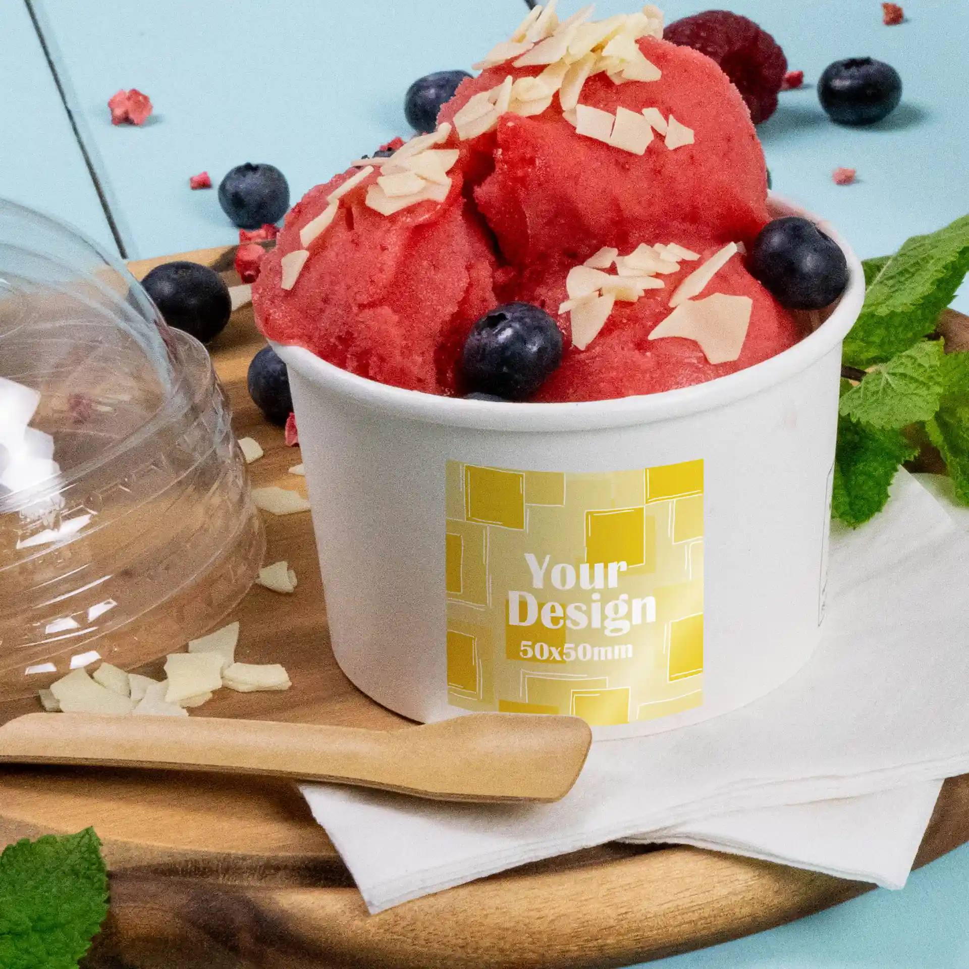 Verre de sorbet rouge garni de flocons d'amande et de myrtilles, avec une étiquette imprimée sur un contenant blanc, posé sur une planche en bois.