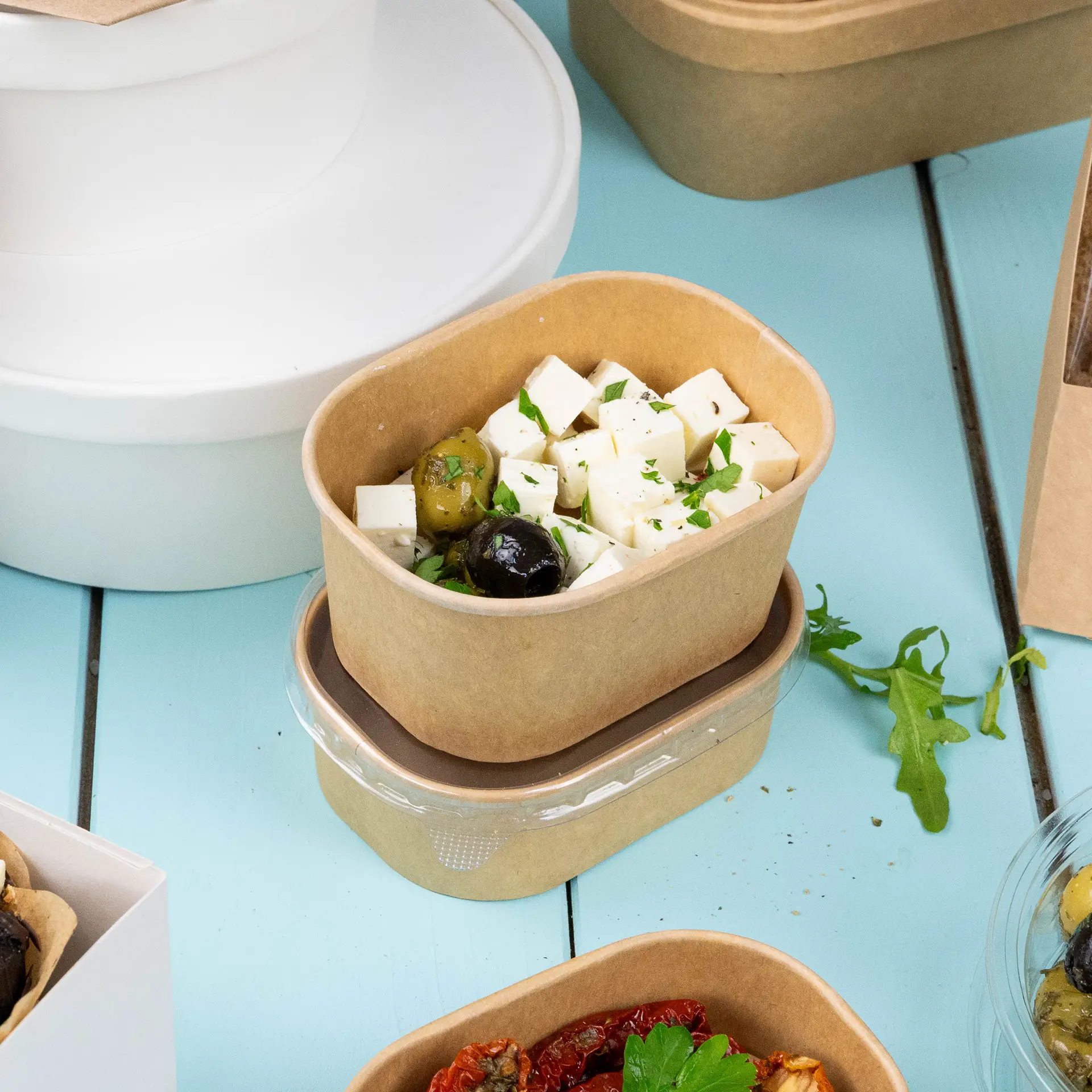 Conteneur alimentaire en kraft rectangulaire rempli de fromage en cubes et d'olives, avec garniture de feuilles; des contenants supplémentaires sont visibles.