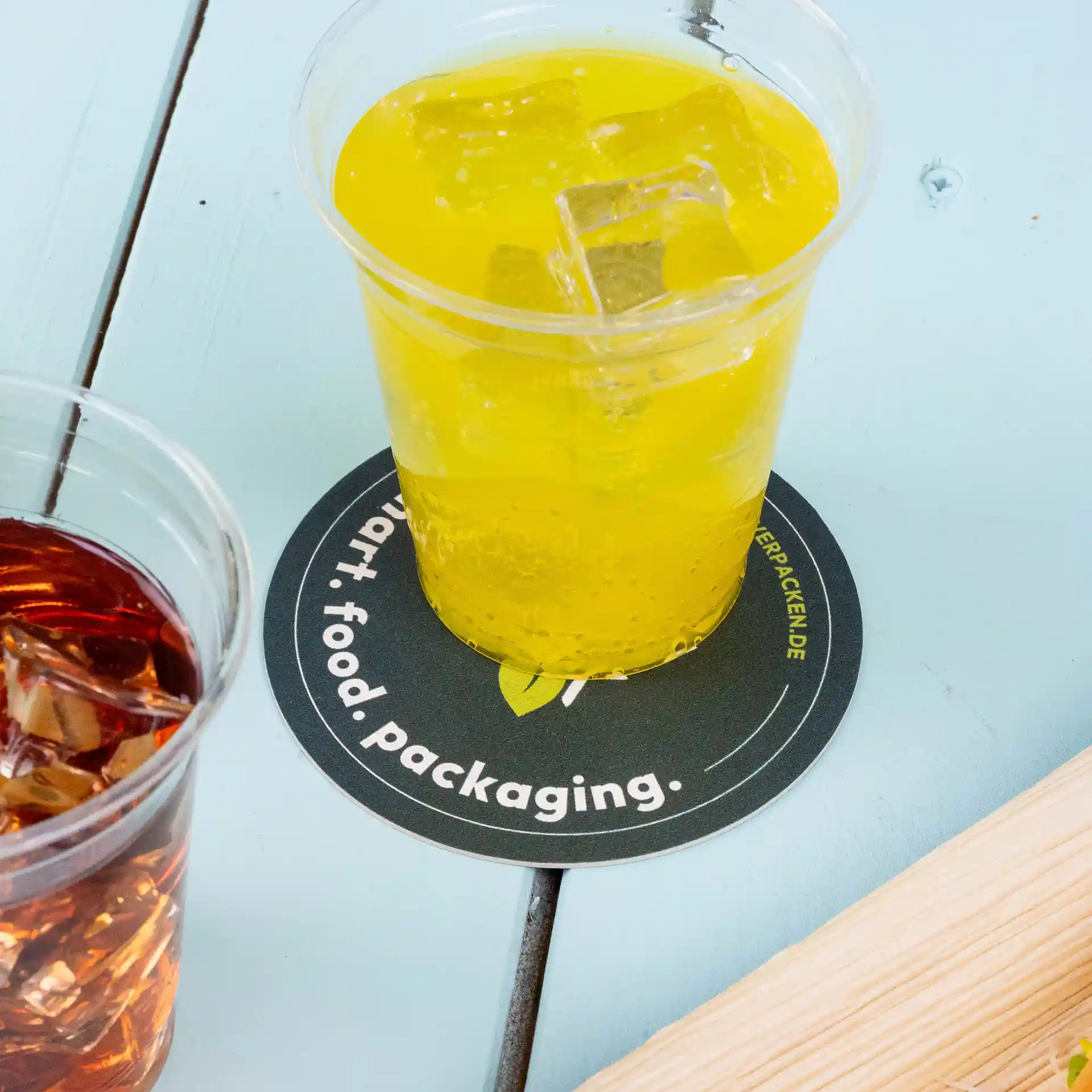 Image d'une boisson jaune avec des glaçons dans un verre transparent sur un sous-verre rond imprimé, avec une boisson brune dans un verre à proximité.