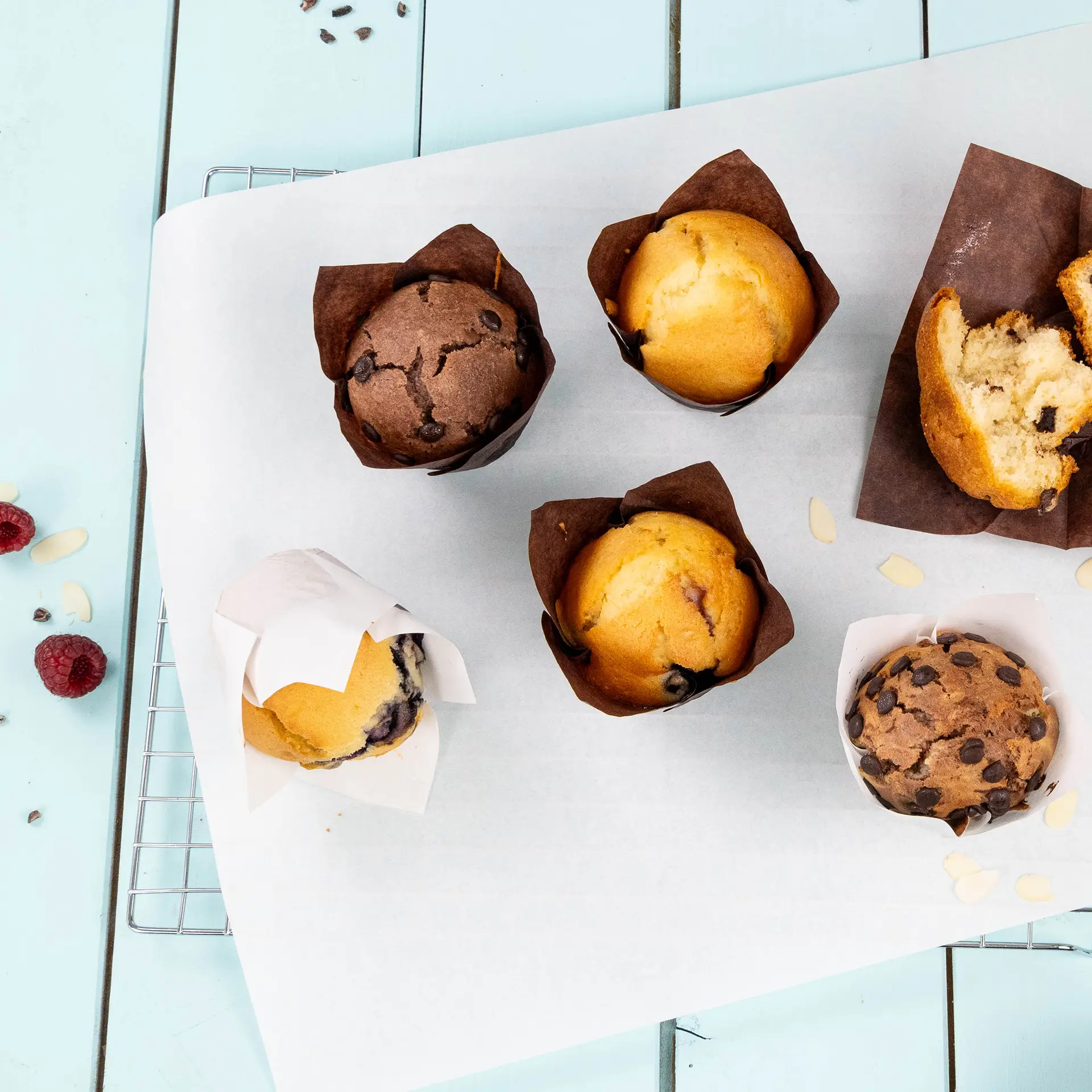 Verschiedene Muffins in braunen Papierbechern auf einer weißen Oberfläche, mit Muffinstücken und verstreuten Mandeln daneben.