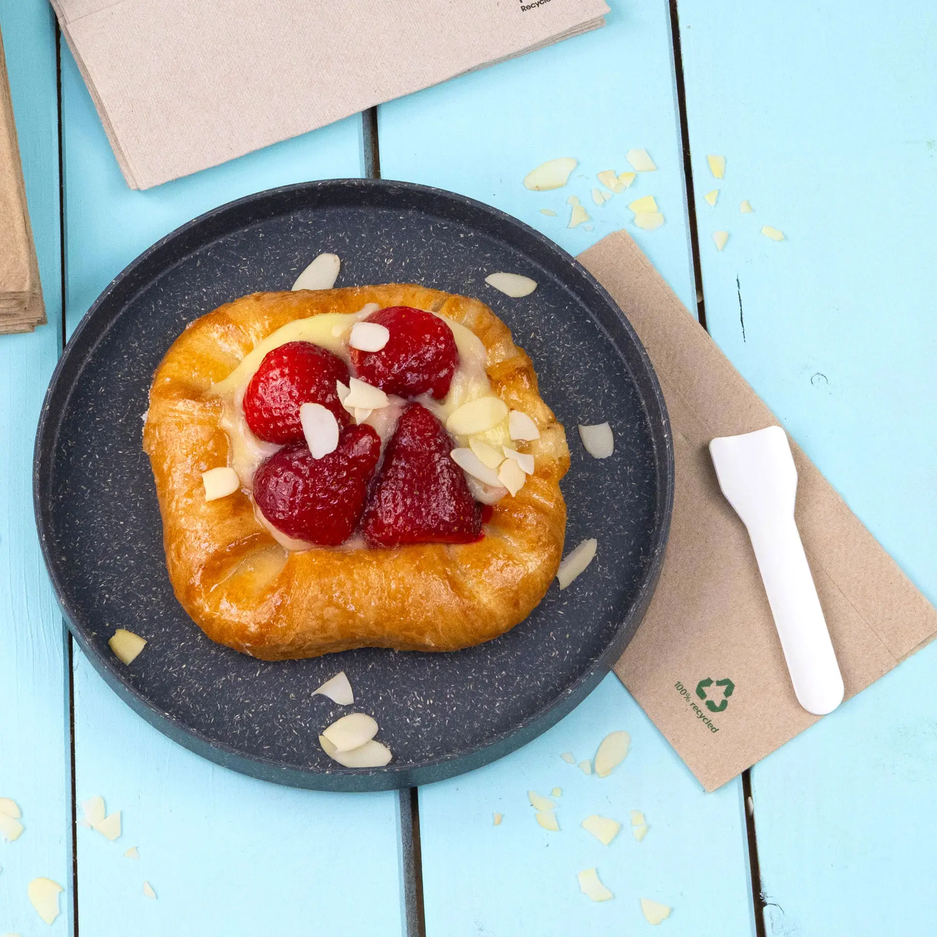 Gebäck mit Erdbeeren und Mandelstücken auf einem dunklen Teller, mit einer Papierserviette und einem Plastikbesteck daneben.