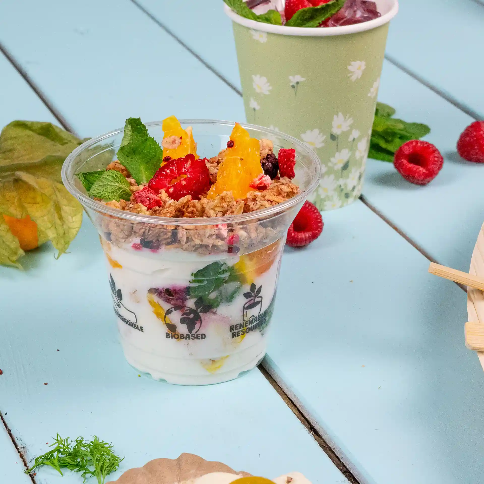 Gobelet transparent rempli de yaourt, granola et fruits, avec des feuilles de menthe sur le dessus, posé sur une surface en bois bleu.