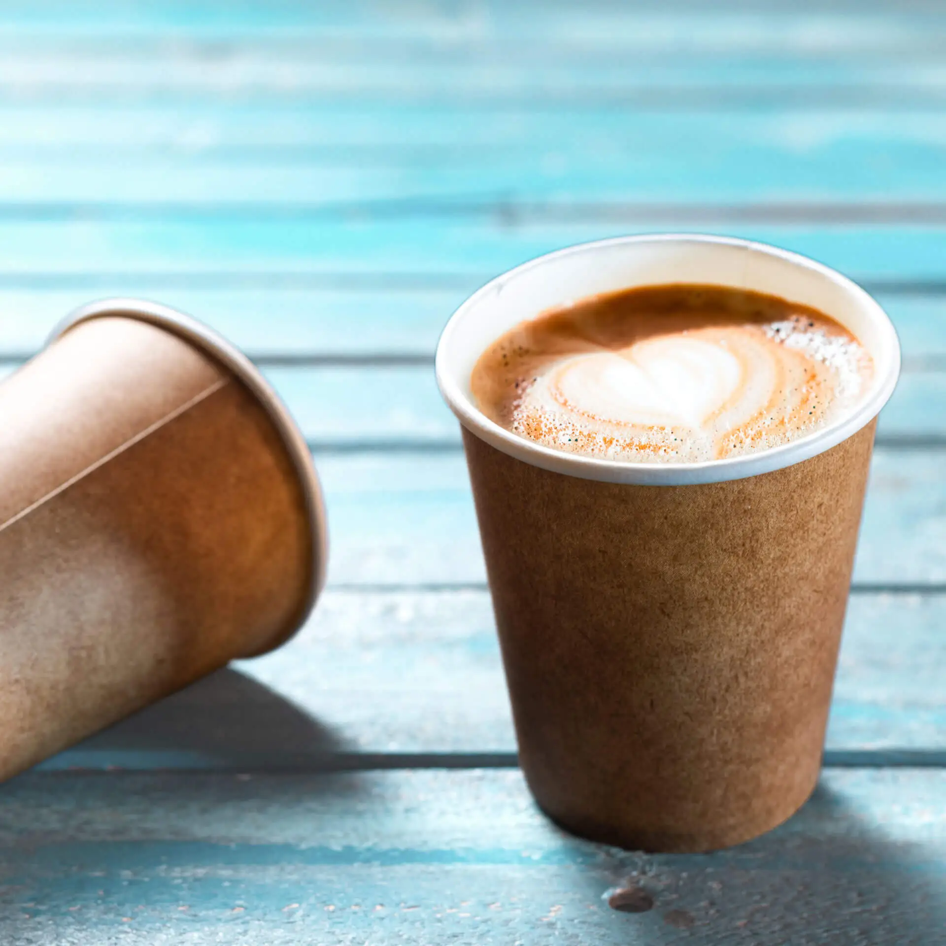 Bicchiere di carta marrone con rivestimento bianco, riempito di caffè, su una superficie in legno blu.
