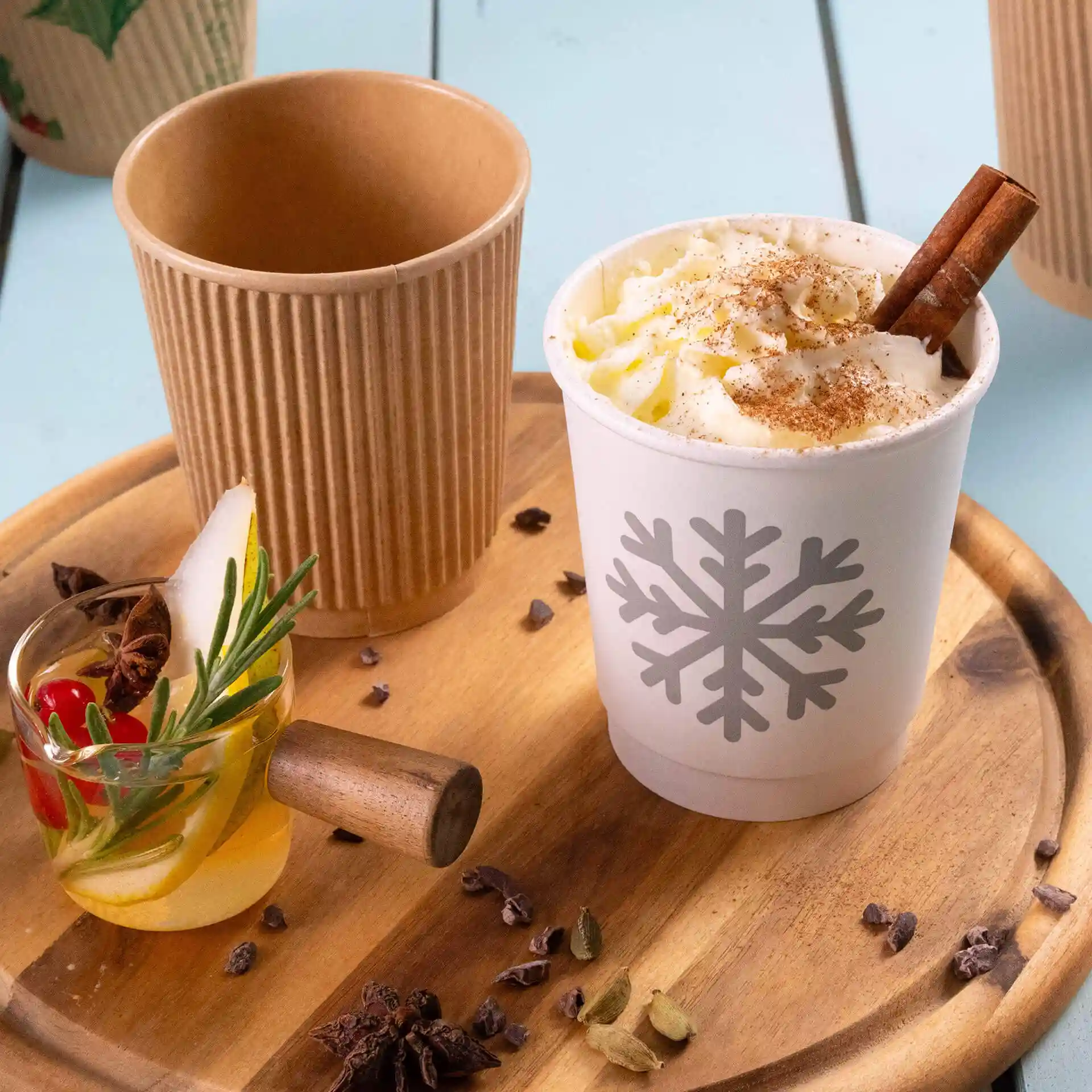 Gobelet blanc avec un motif de flocon de neige, rempli de crème fouettée et d'un bâton de cannelle, à côté d'un gobelet kraft et d'une boisson garnie.