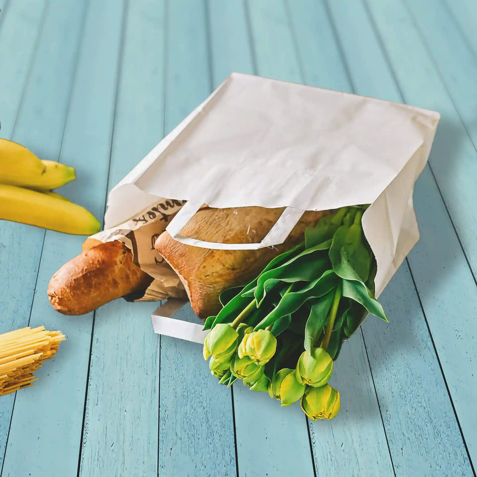 Weißer Kraftpapier-Tragetasche mit breitem Boden, gefüllt mit Brot, Tulpen und umgeben von Bananen und Spaghetti.