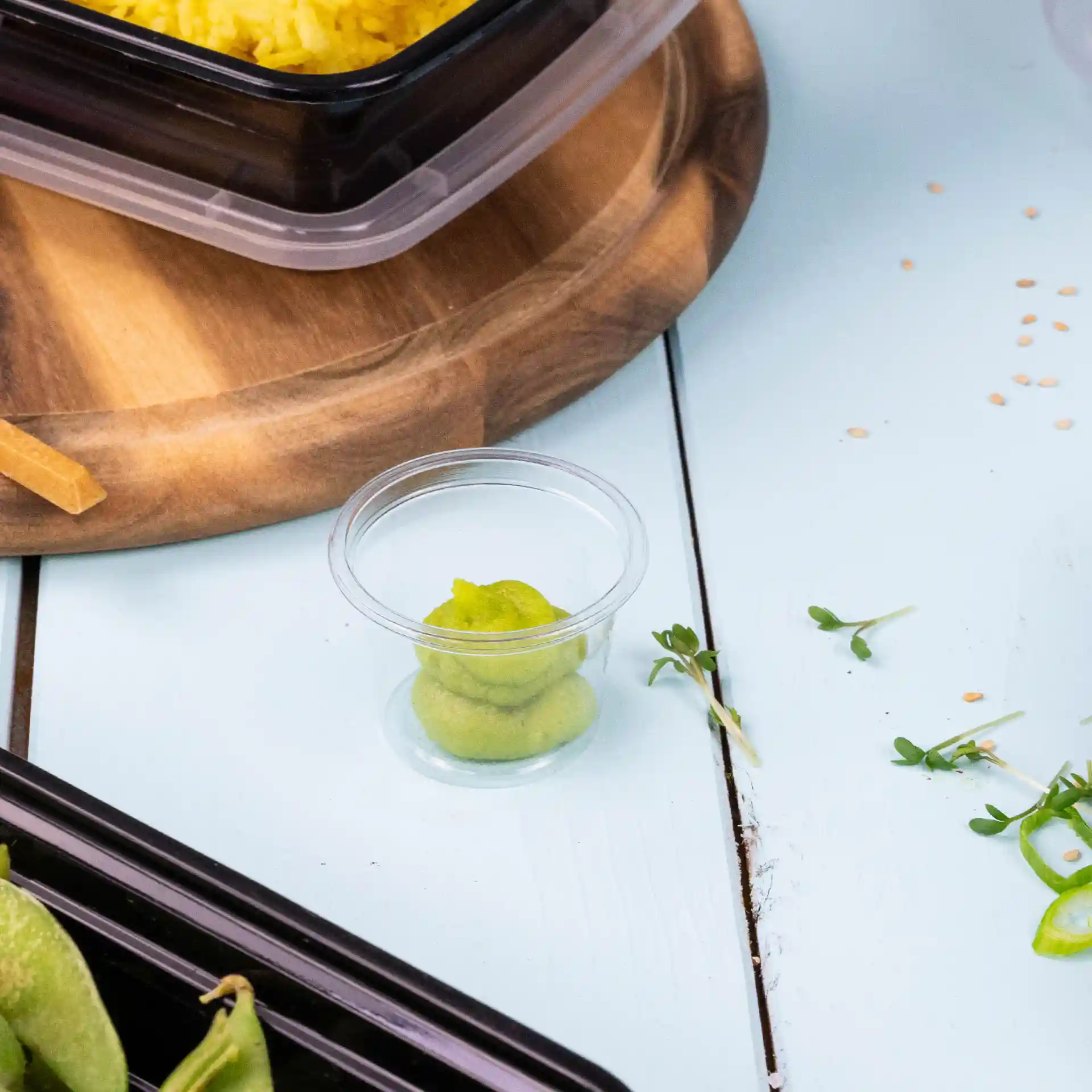 Gobelet en PLA transparent avec condiment vert à l'intérieur, placé sur une surface bleu clair à côté d'une planche en bois.