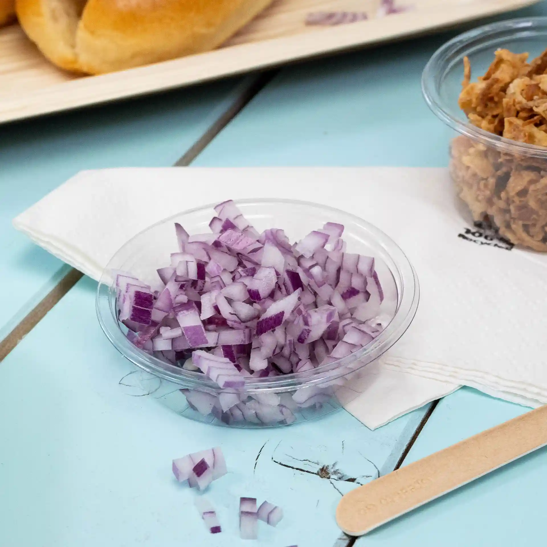 Klarer runder Becher mit fein gewürfelten roten Zwiebeln auf einer blauen Oberfläche, mit einer Serviette und einem Holzstab in der Nähe.