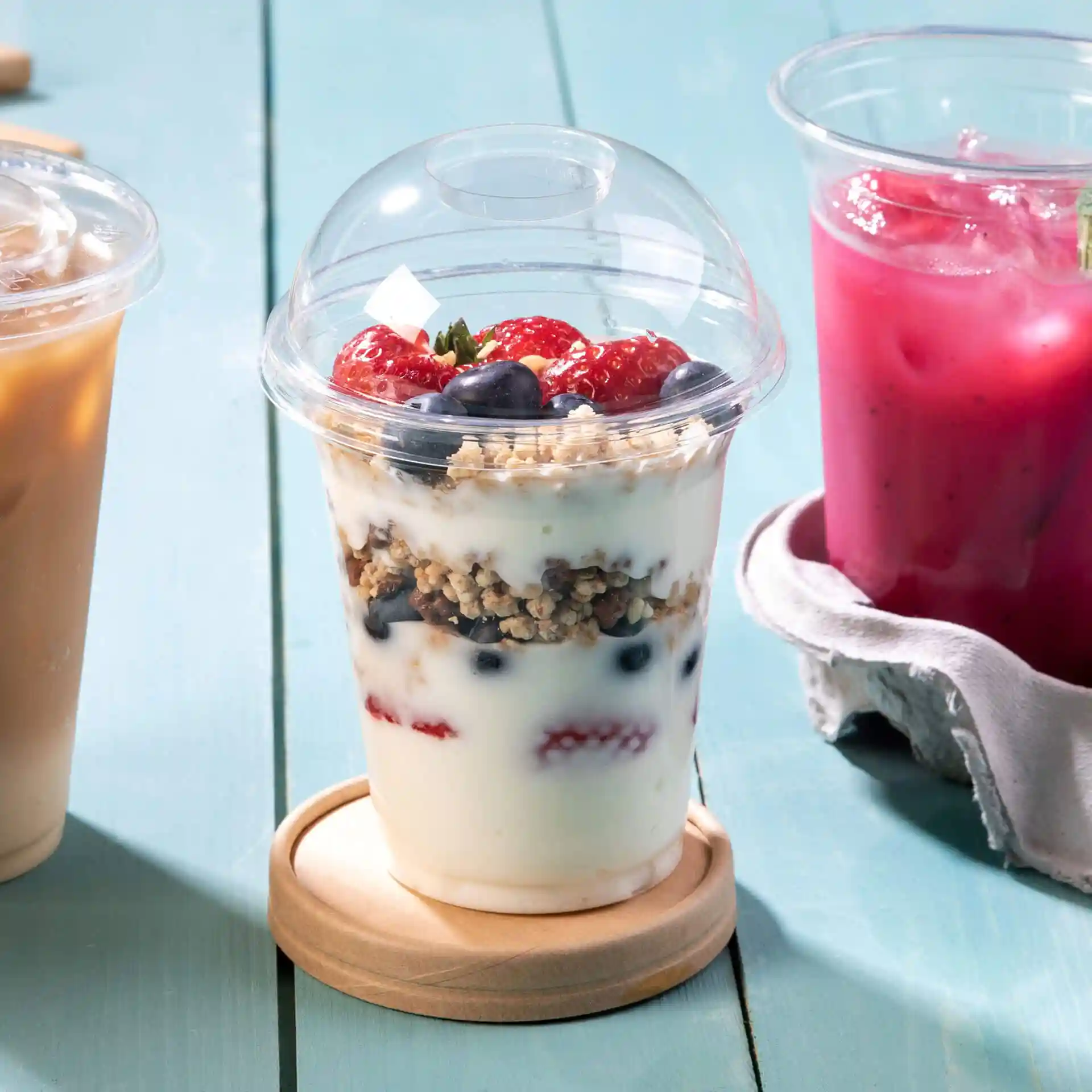 Coperchio a cupola trasparente per un bicchiere, con granola, yogurt e frutti di bosco, posato su un sottobicchiere di legno.