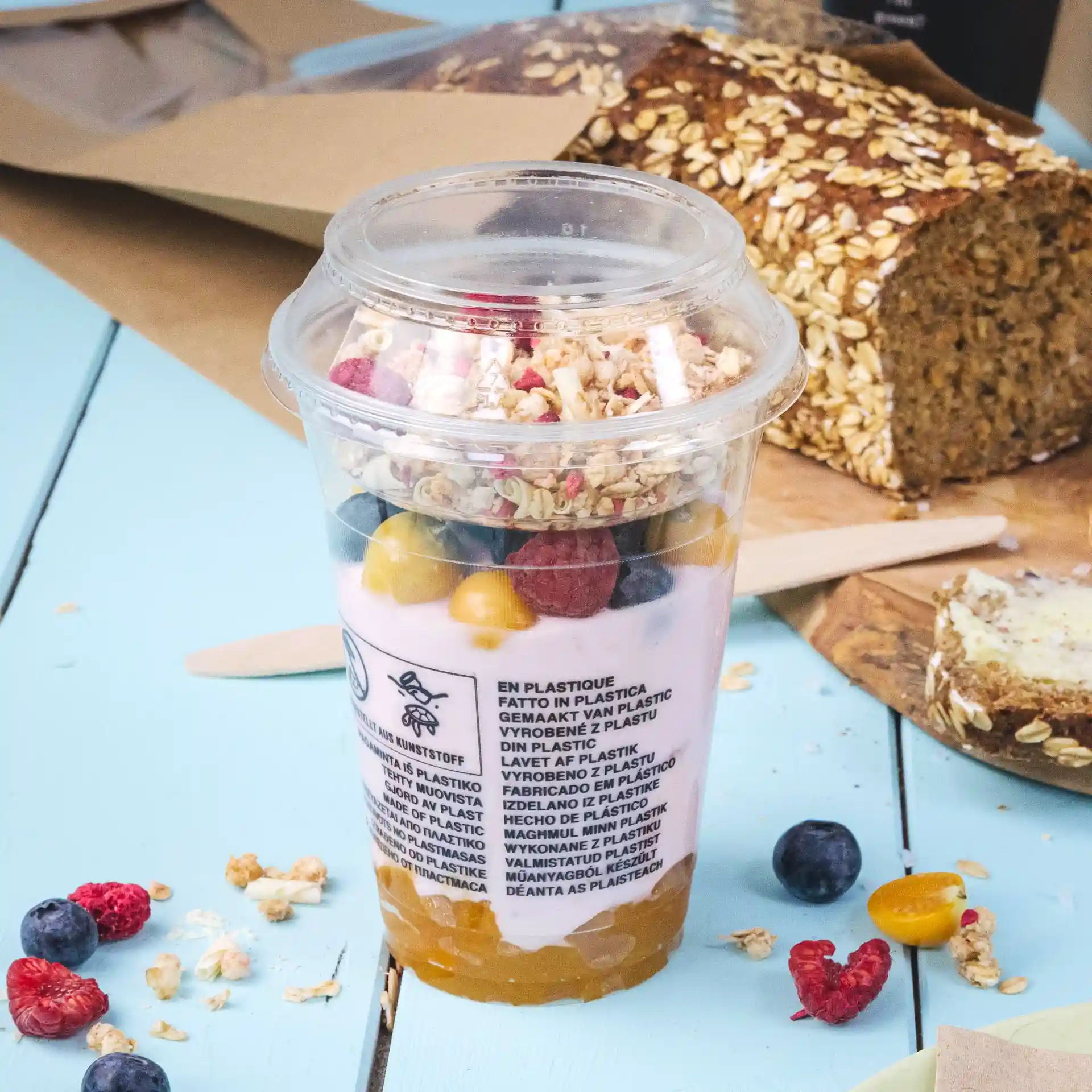 Bicchiere smoothie PLA trasparente da 12 oz riempito di yogurt, granola e frutta mista, posato su una superficie blu.