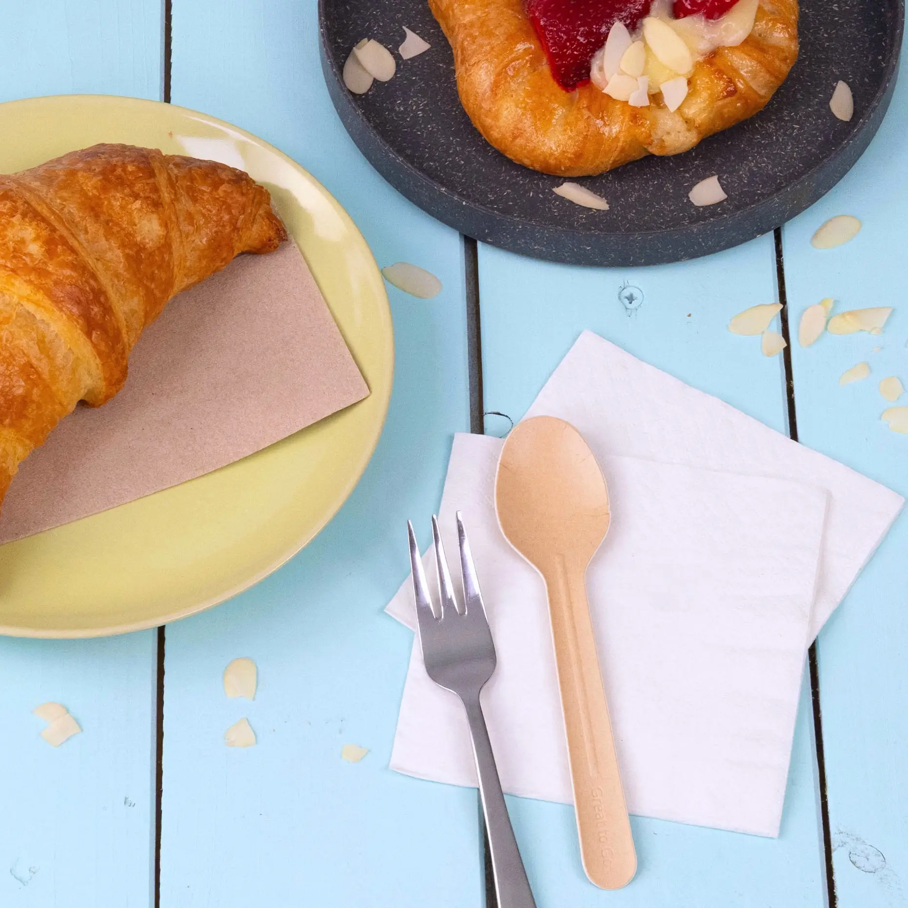 Bild eines Croissants auf einem gelben Teller, einem Gebäck auf einem dunklen Teller und einem Papierspoon neben einer Gabel auf Servietten.