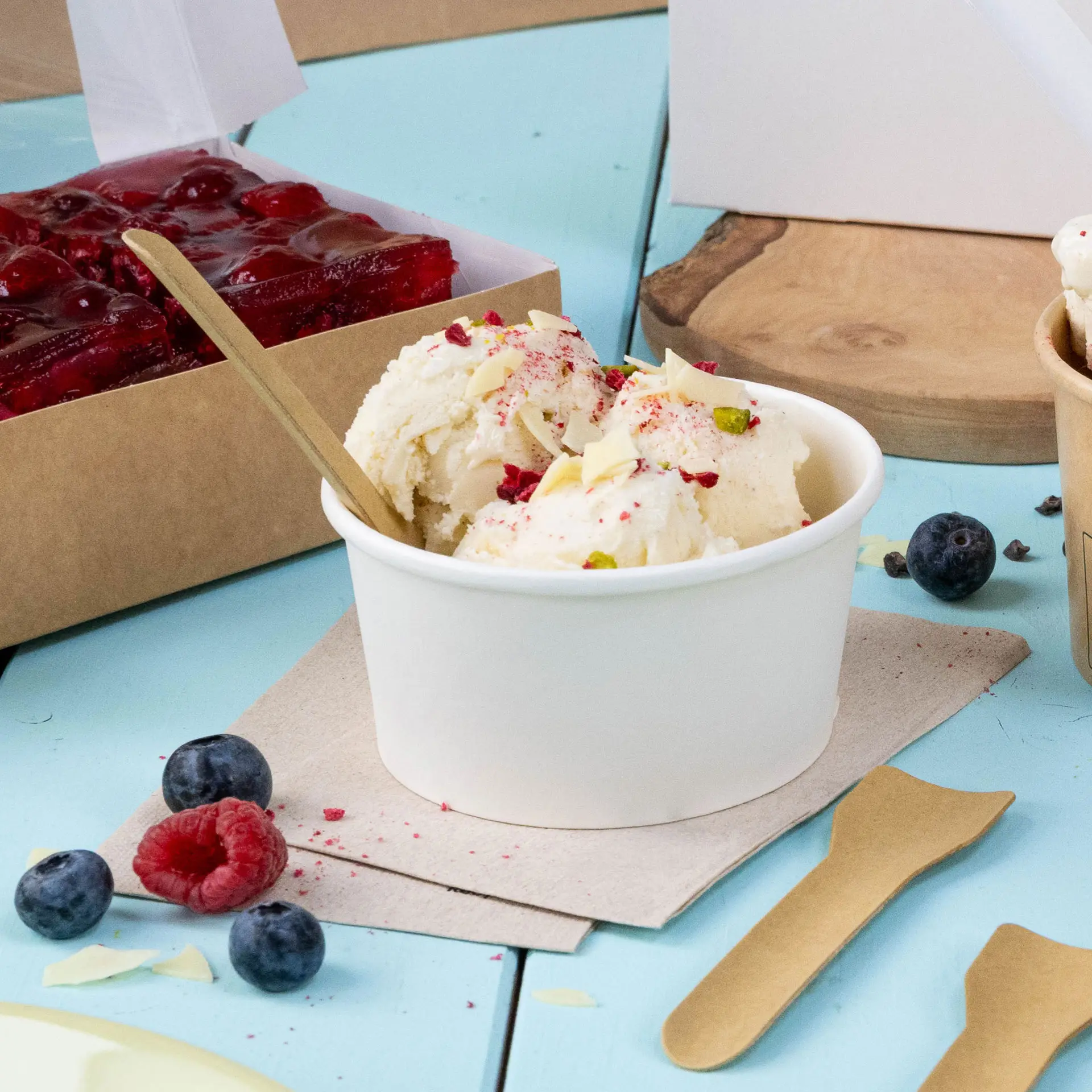 Becher mit Eis, garniert mit Mandelscheiben und Himbeerstücken, daneben Heidelbeeren und Himbeeren auf blauer Fläche.