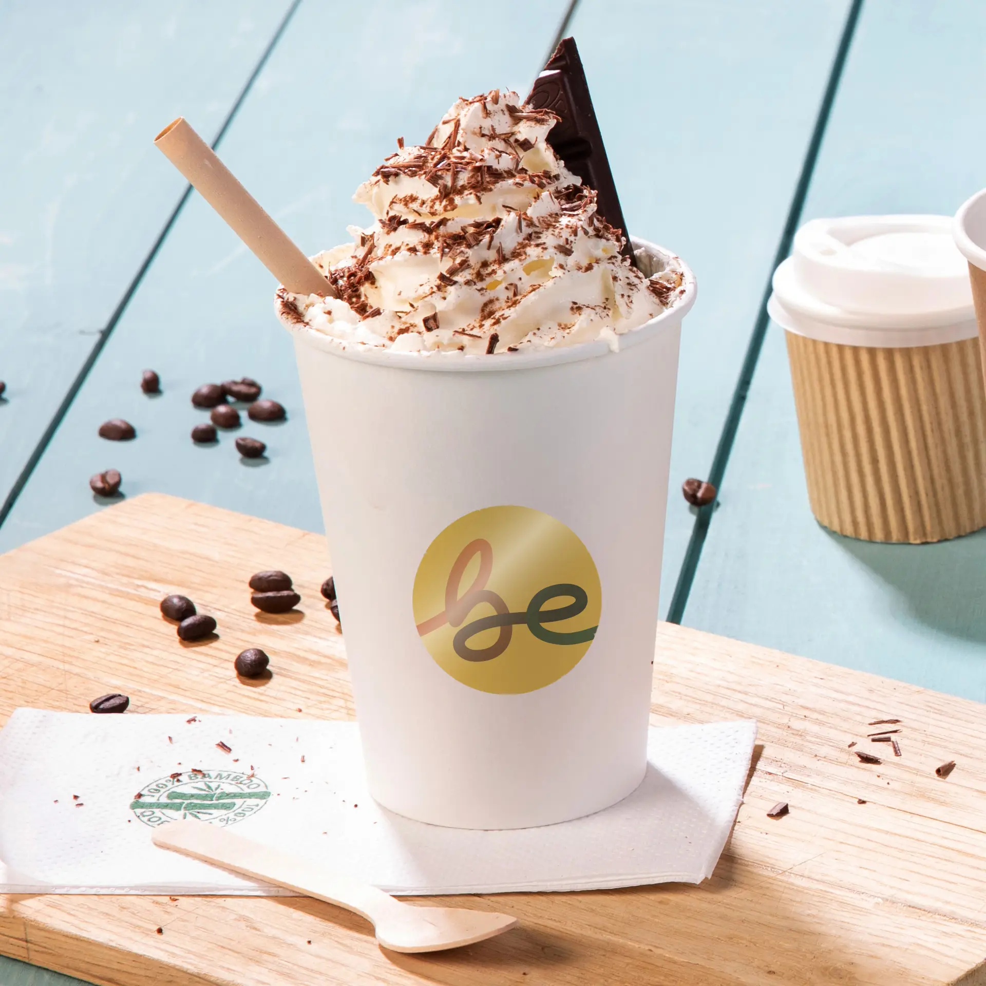 Tasse avec de la crème fouettée, des flocons de chocolat, un morceau de chocolat et une paille en papier, posée sur une surface en bois avec des grains de café.