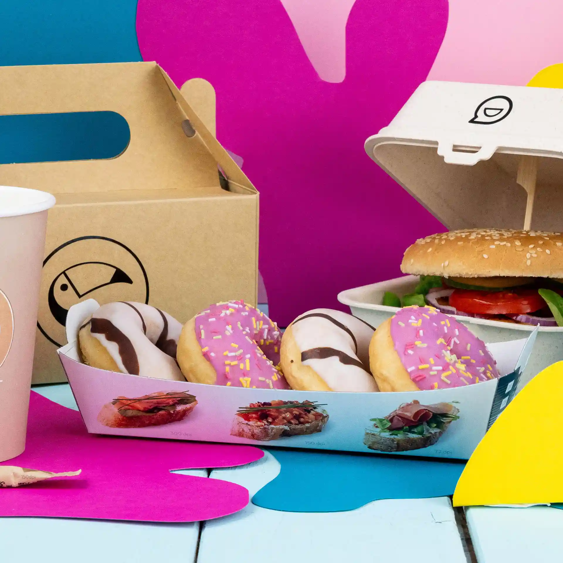 Plateau rectangulaire blanc avec quatre donuts, un burger dans un contenant et une tasse rose sur un fond coloré.