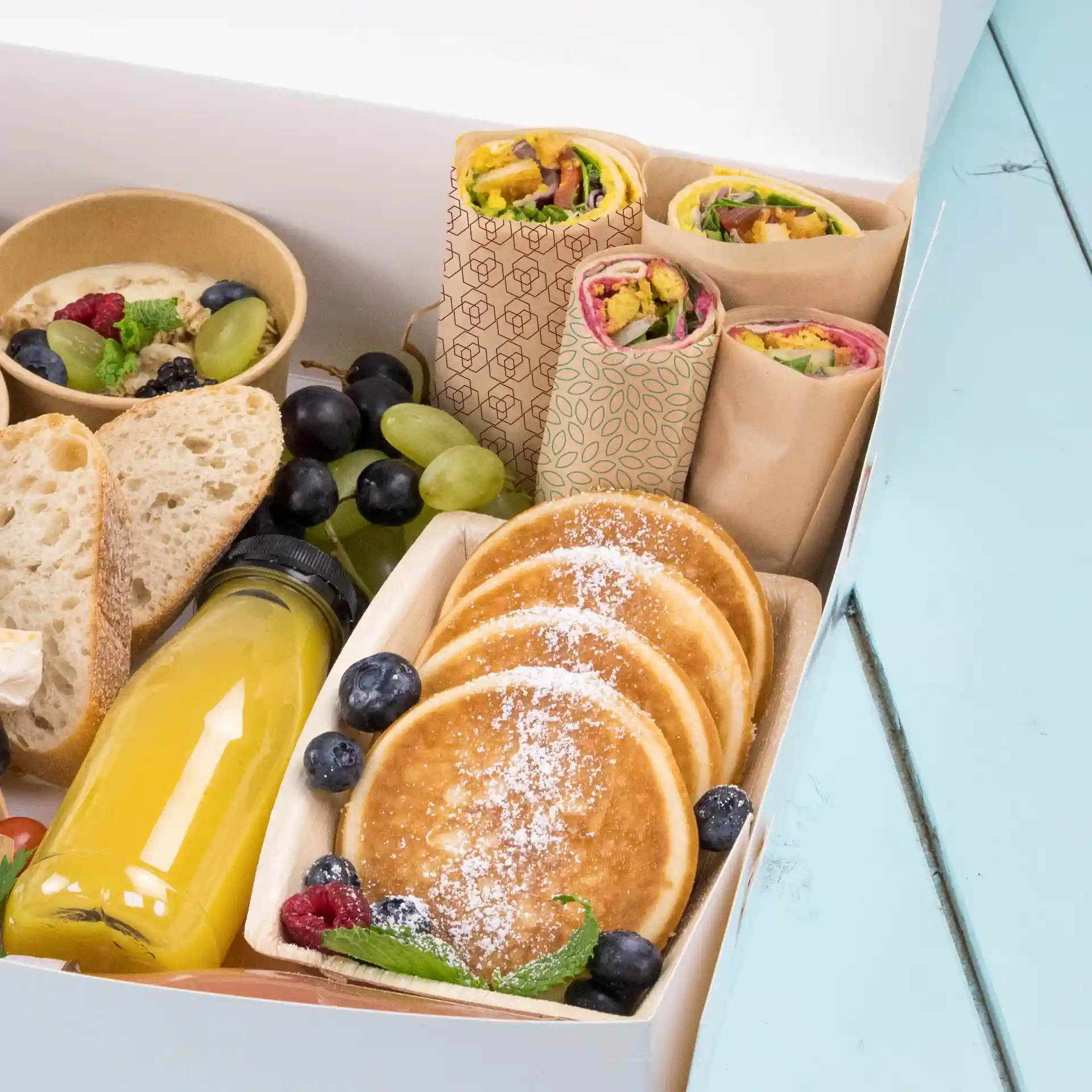 Assortiment d'aliments comprenant des wraps, des crêpes, des fruits et une boisson dans une boîte sur une surface bleue.