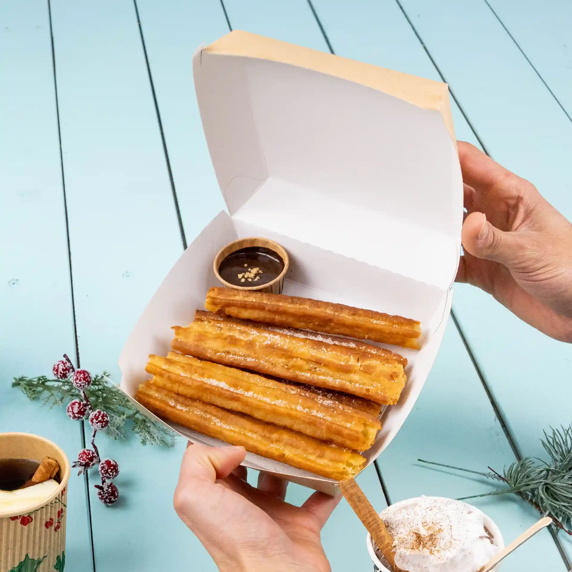 Scatola da asporto con diversi churros e un piccolo bicchiere di salsa al cioccolato, su una superficie in legno blu chiaro.