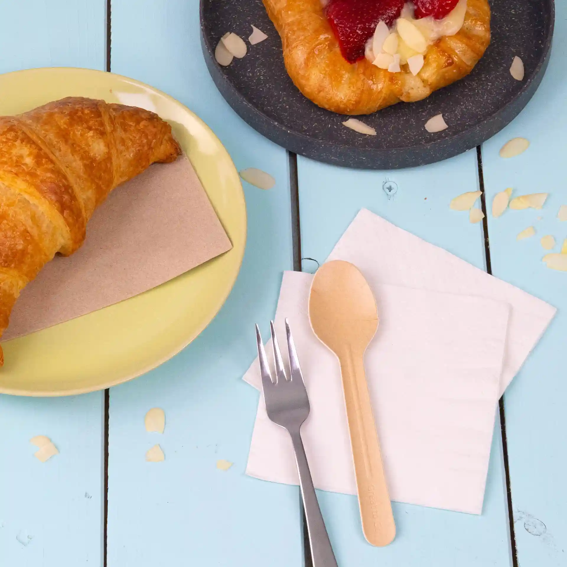 Immagine di un croissant su un piatto giallo, un dessert su un piatto scuro, un cucchiaio di legno, una forchetta d'argento e tovaglioli bianchi.