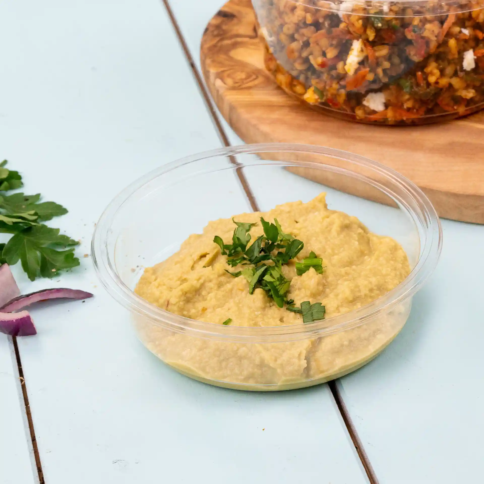 Contenitore deli trasparente rotondo con hummus guarnito con prezzemolo tritato su una superficie azzurra chiara.