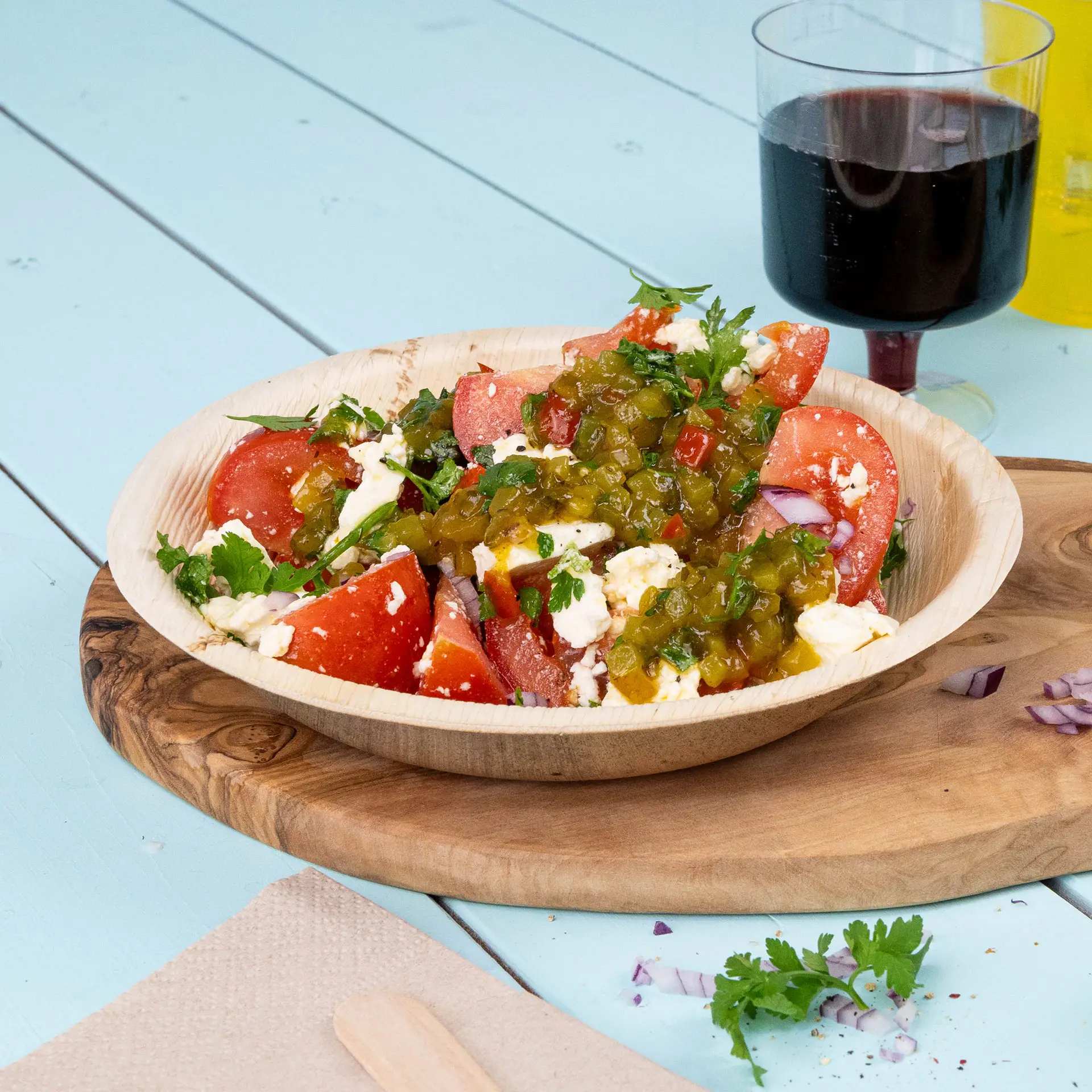 Une assiette ronde en feuille de palmier remplie d'une salade de tomates, herbes et jalapeño, posée sur une planche en bois avec une boisson.