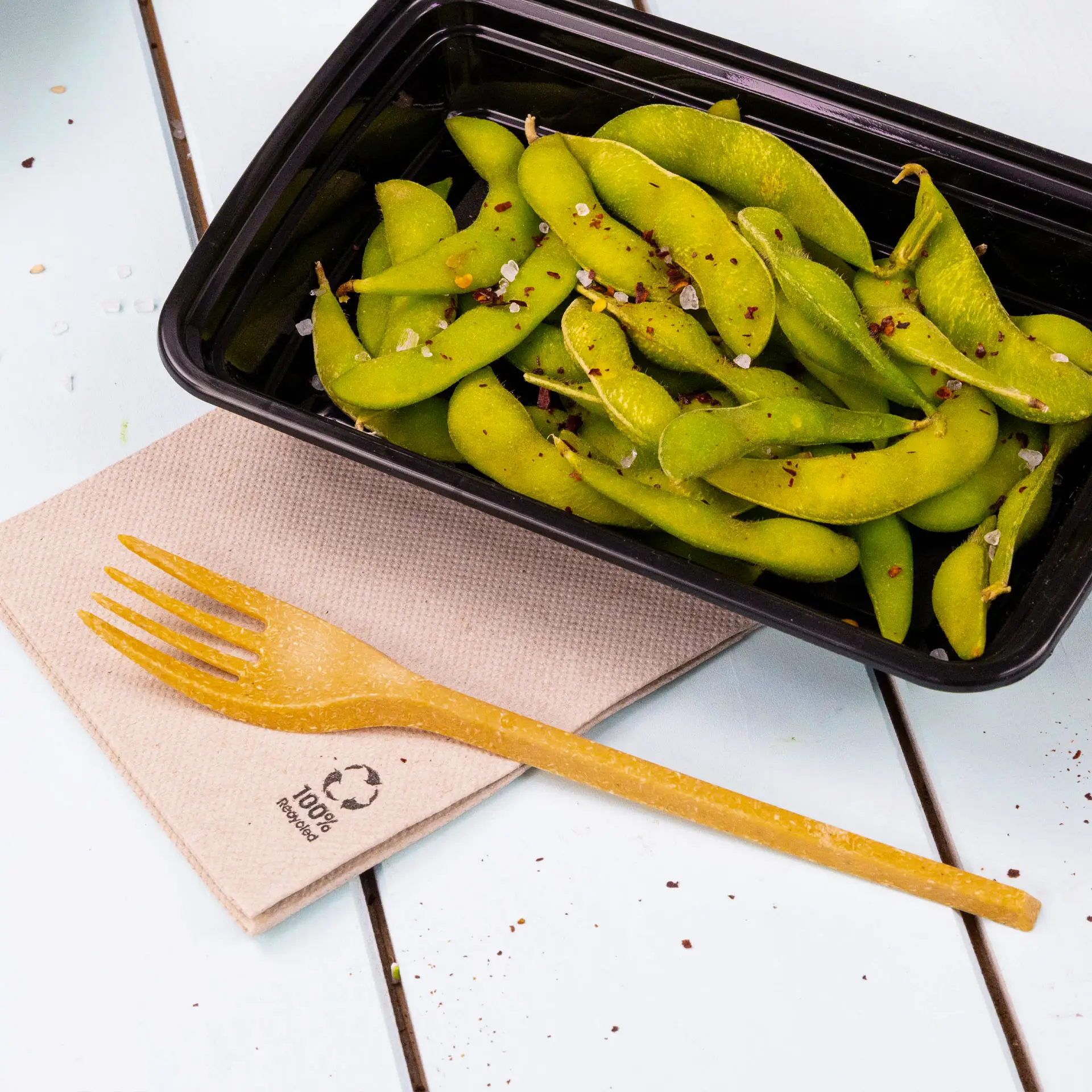 Image de haricots edamame frais dans un contenant noir, accompagnés d'une fourchette biodégradable sur une serviette en papier recyclé.