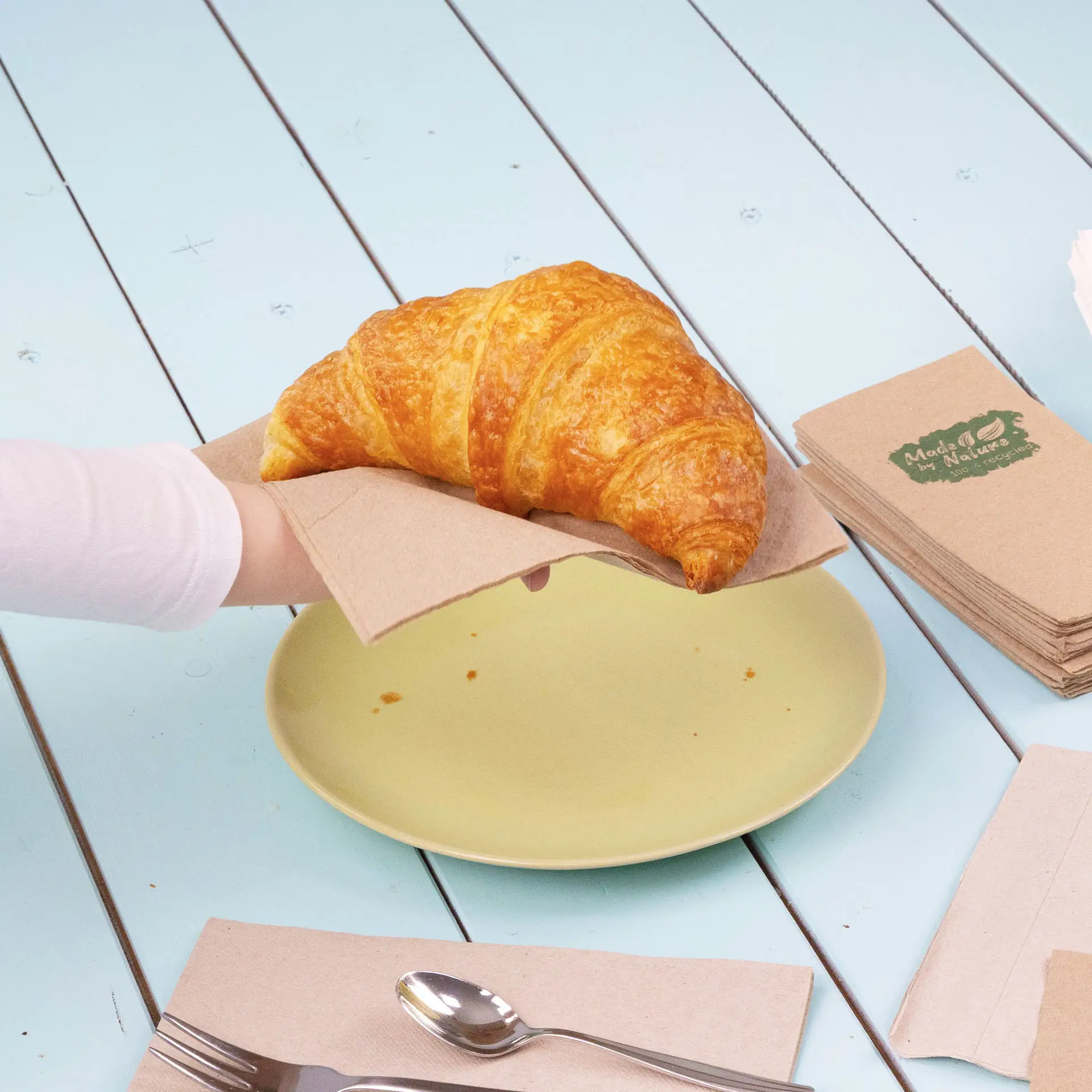 Eine Hand hält ein Croissant auf einer Papierserviette über einem gelben Teller, mit weiteren Servietten und Besteck in der Nähe.