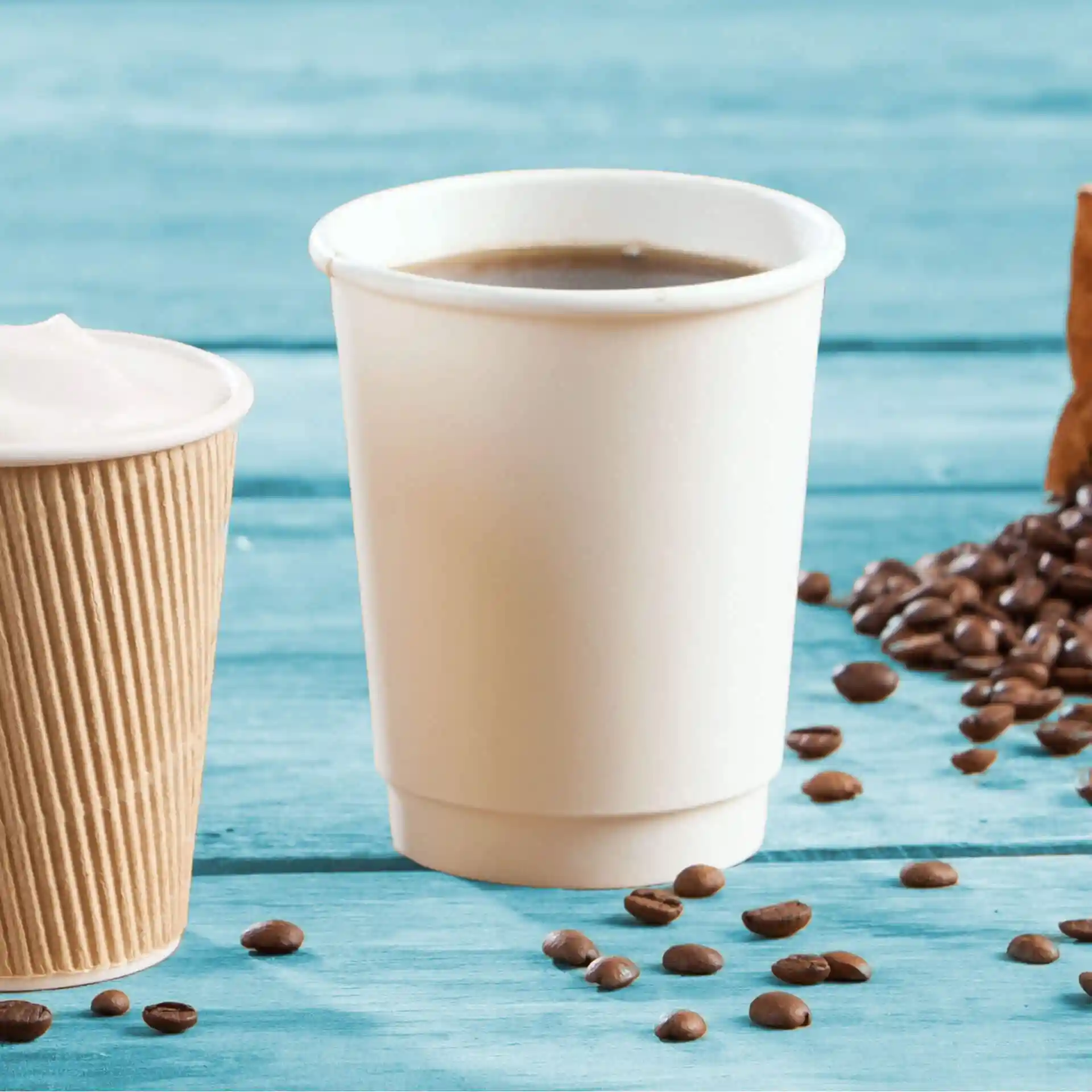 Bicchiere di cartone bianco a doppia parete con caffè, accanto a una tazza marrone rigata e chicchi di caffè sparsi.