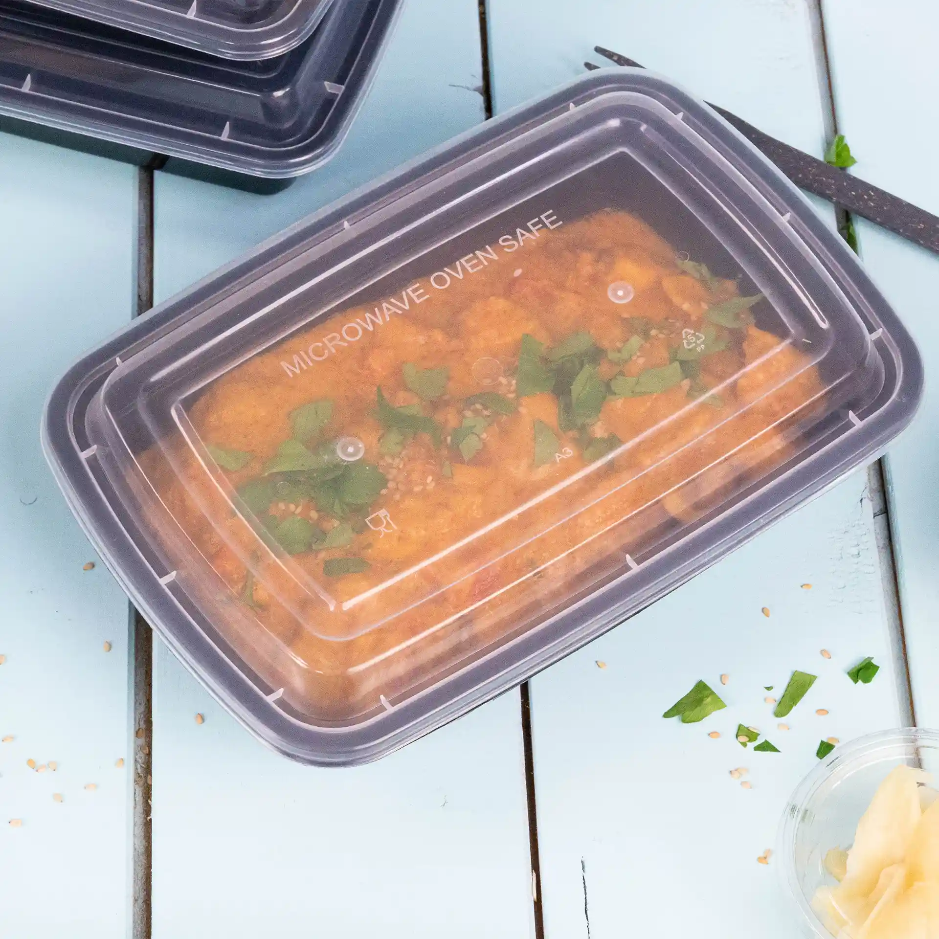 Contenitore rettangolare trasparente con coperchio riutilizzabile, riempito di cibo e guarnito con erbe verdi, su una superficie blu.