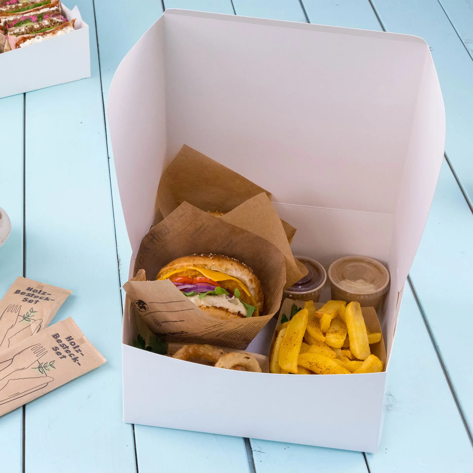 Boîte à gâteau en carton blanc contenant un sandwich, des frites et deux contenants de sauce, avec un couvercle plié.