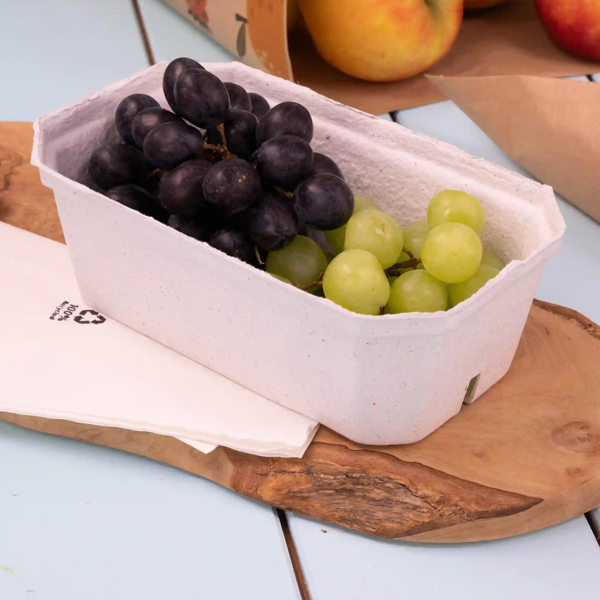 Conteneur avec des raisins noirs et verts dans un barquette en pulpe de bois blanc sur une surface en bois avec des serviettes à proximité.