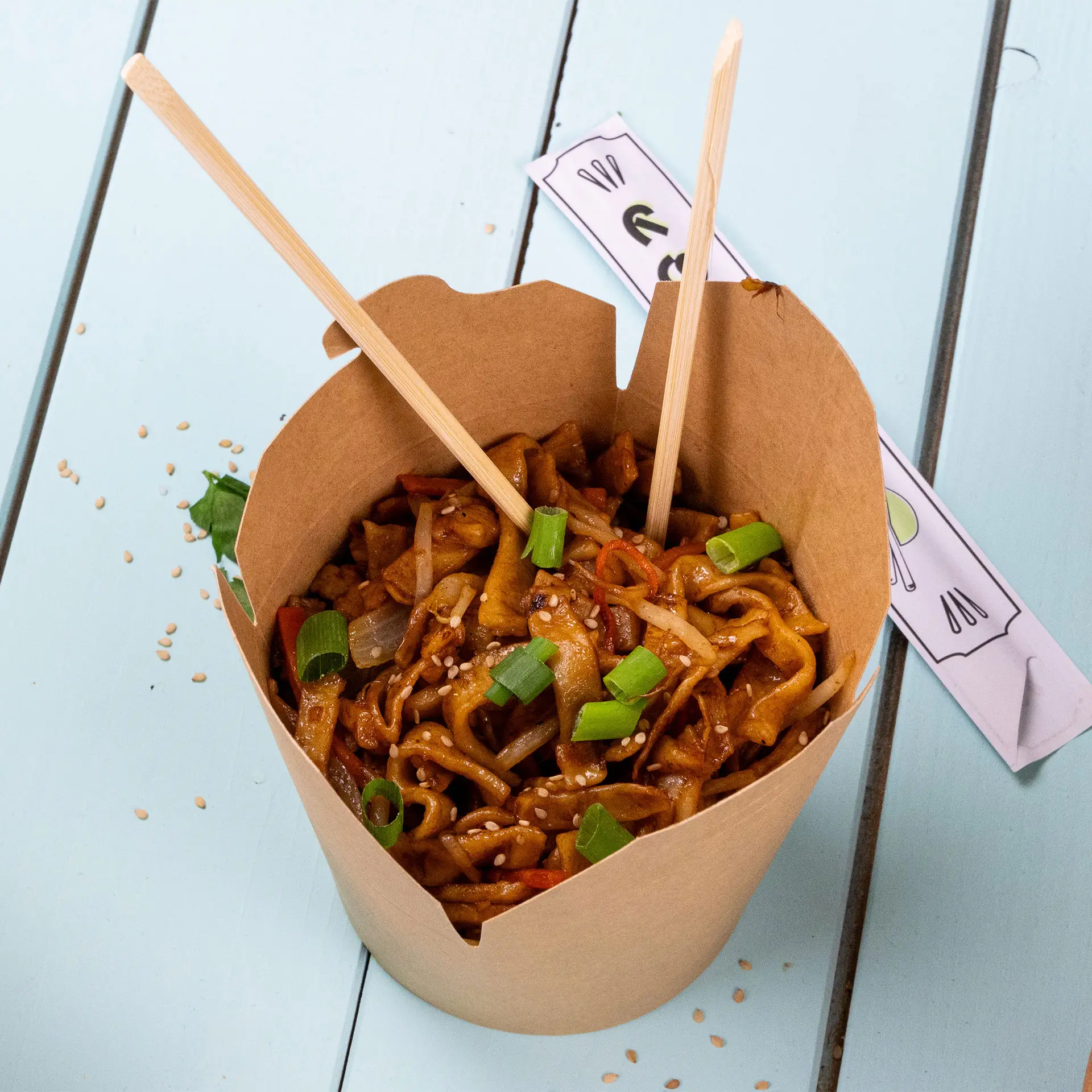 Boîte à nouilles en kraft marron remplie de nouilles frites, garnie d'oignons verts et de graines de sésame, avec des baguettes.
